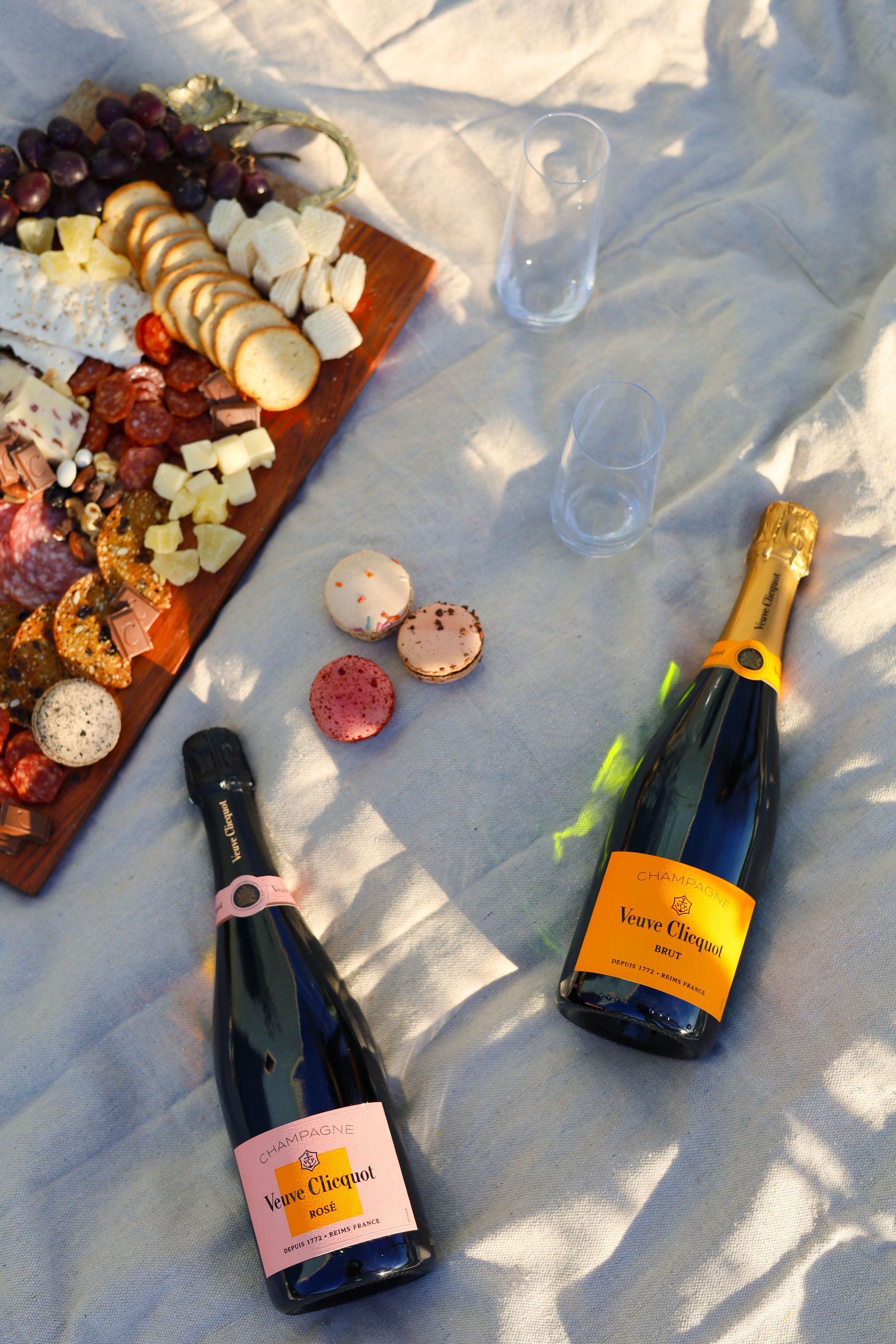 Two champagne bottles on a picnic blanket beside a charcuterie board and scattered macarons.