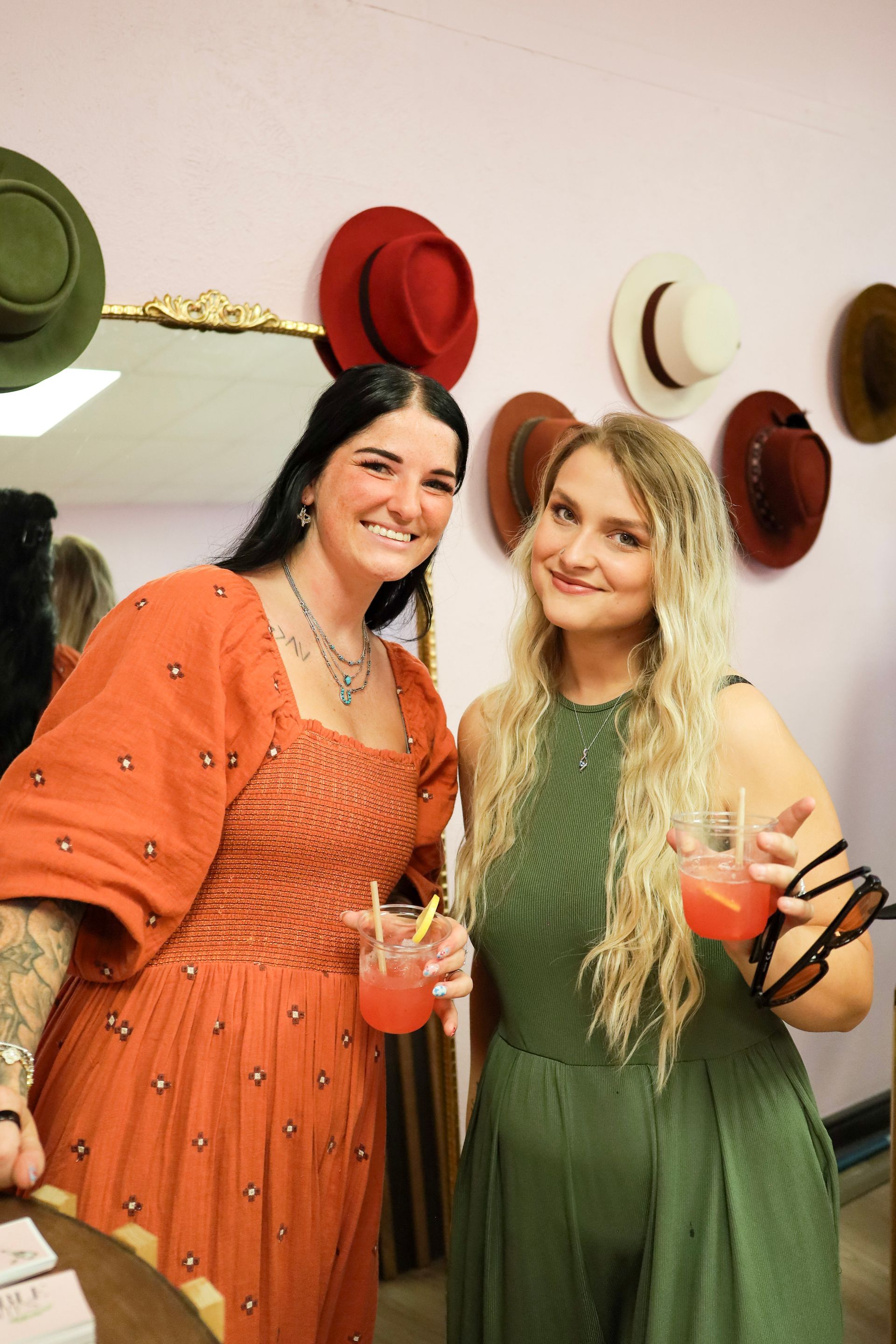 Two women holding orange drinks, smiling in front of a wall with colorful hats.