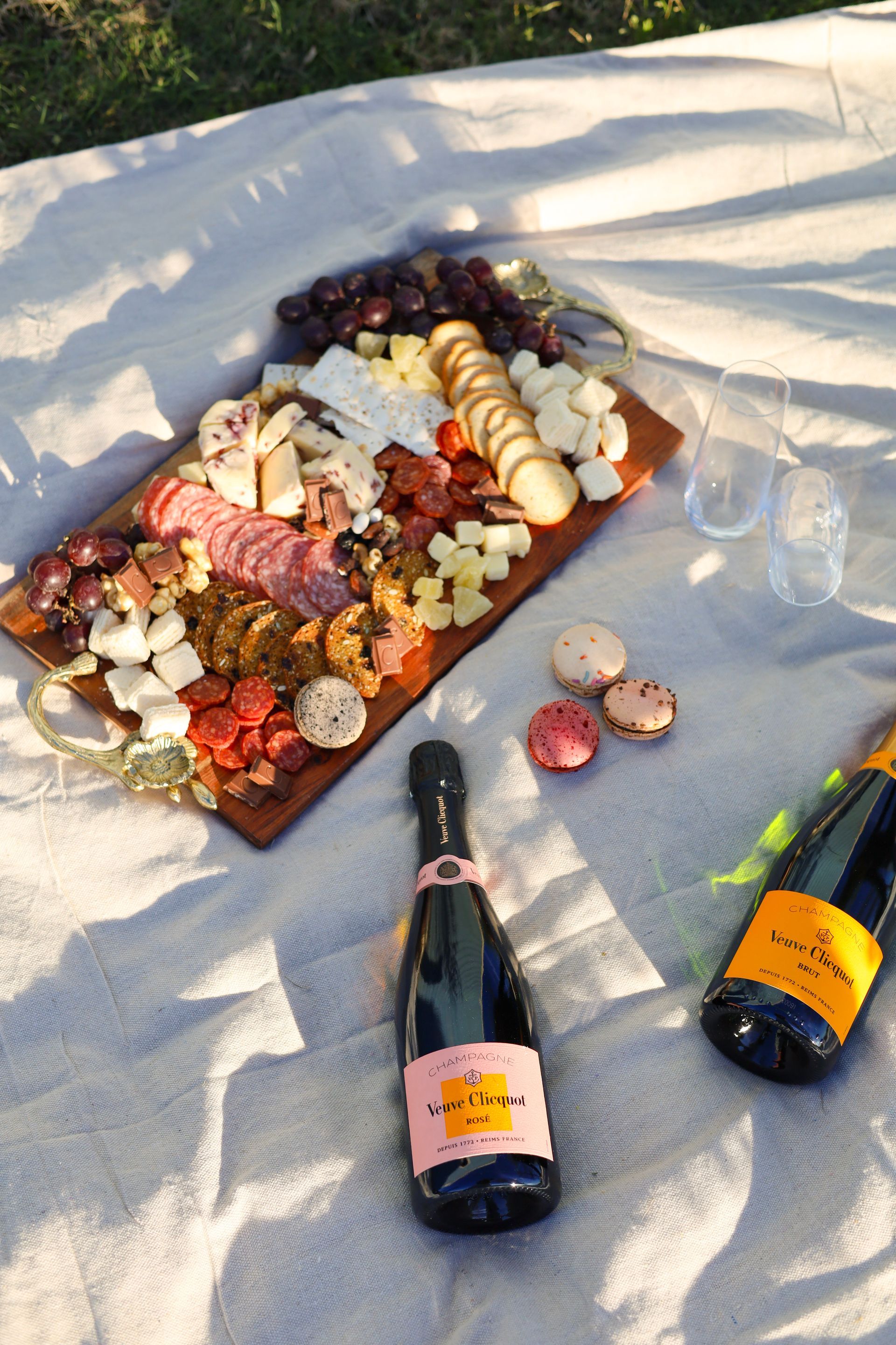 Picnic charcuterie board on a blanket with cheese, fruit, crackers, and two bottles of champagne.