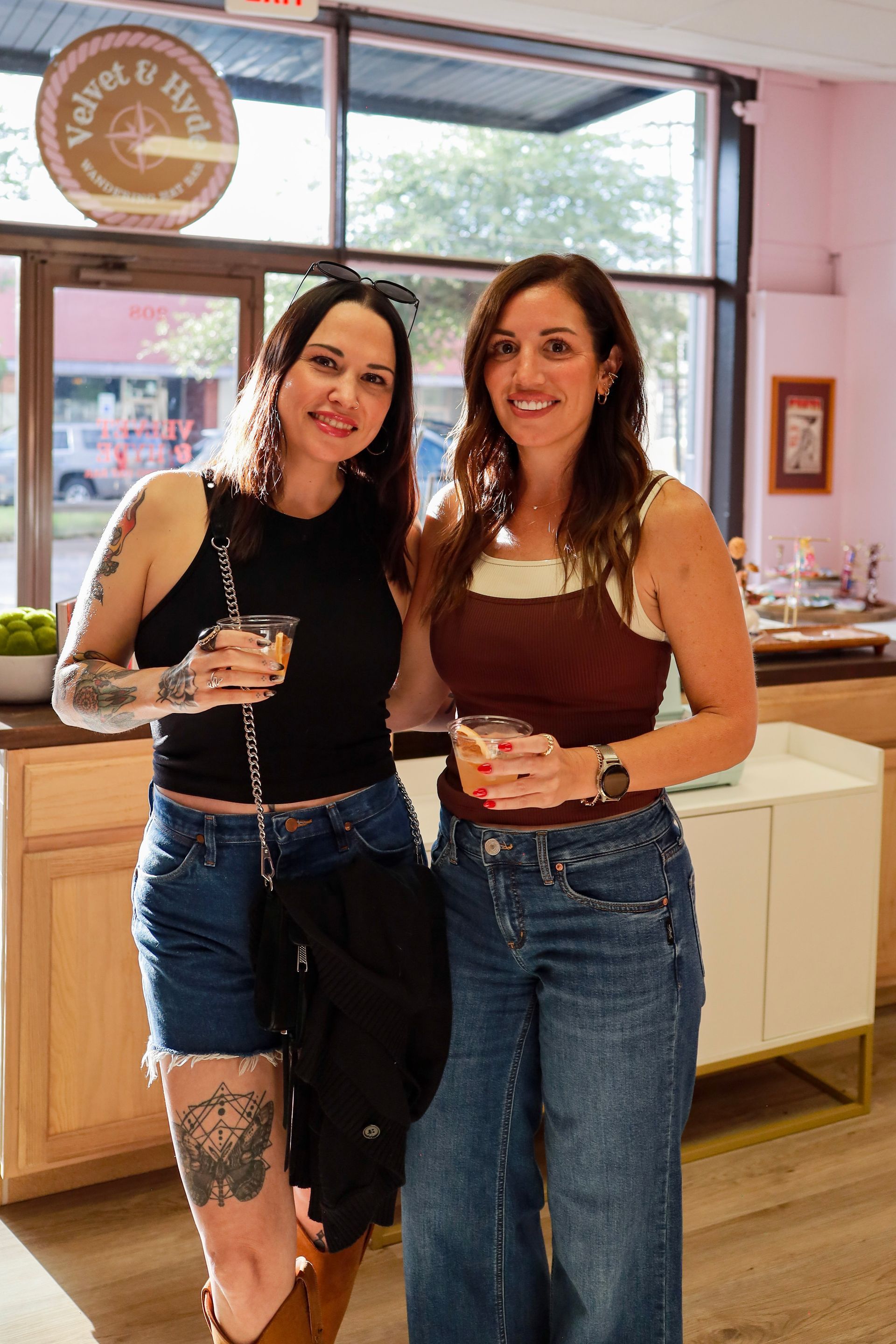 Two women smiling and holding drinks in a bright cafe, standing by a window.
