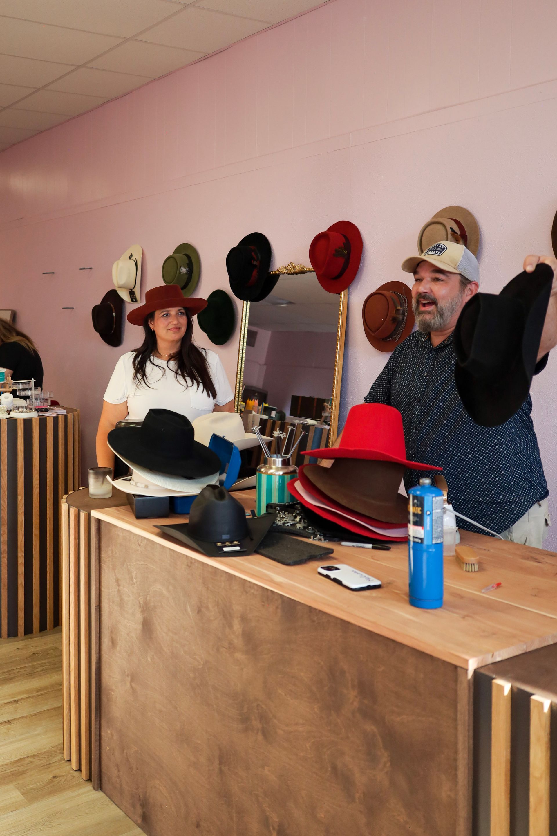 Two people behind a shop counter with hats and accessories displayed on shelves and the counter