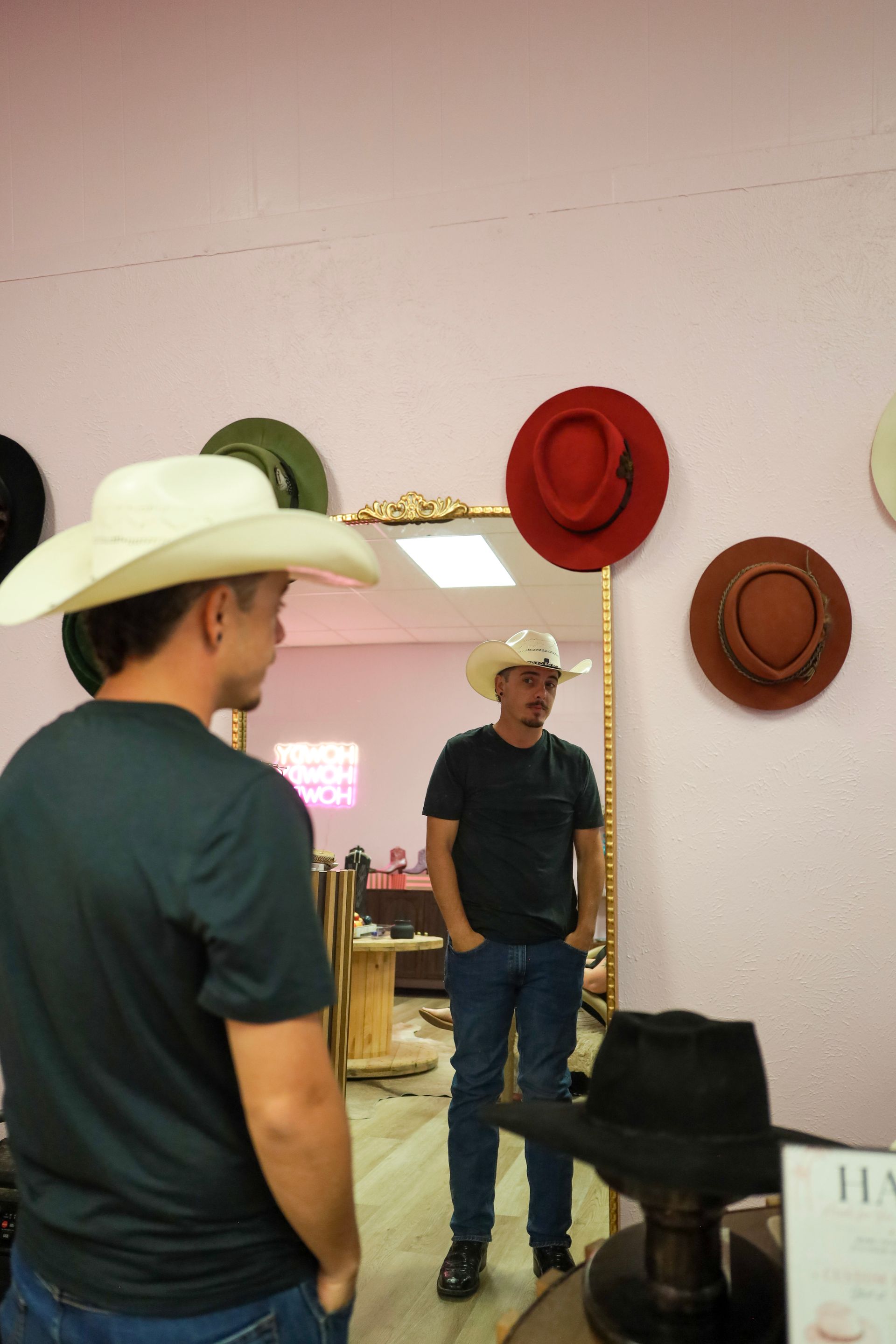 Person in a white cowboy hat looks at their reflection in a mirror beside hanging hats.