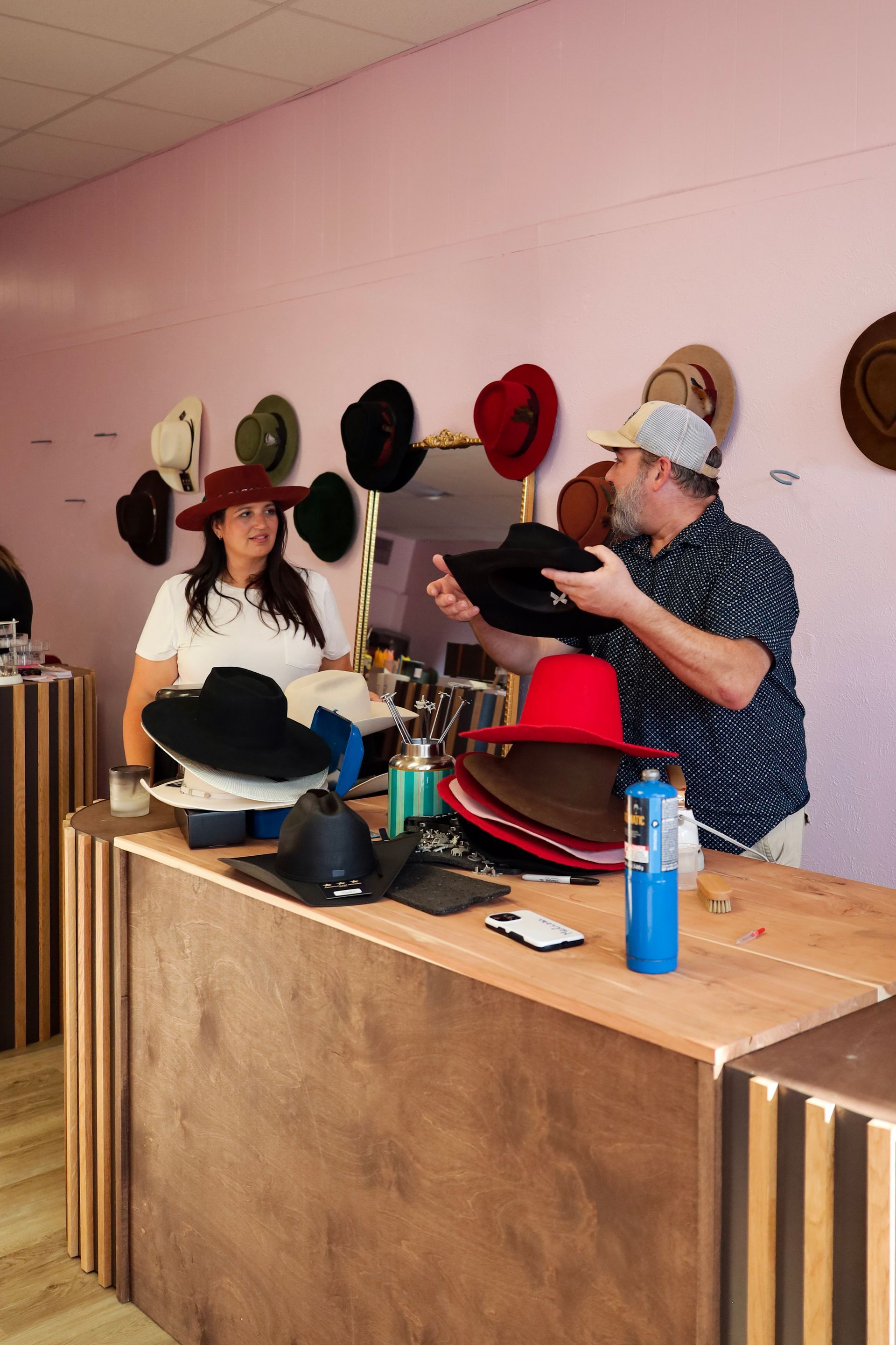 Two people arranging hats behind a wooden counter in a pink-walled shop