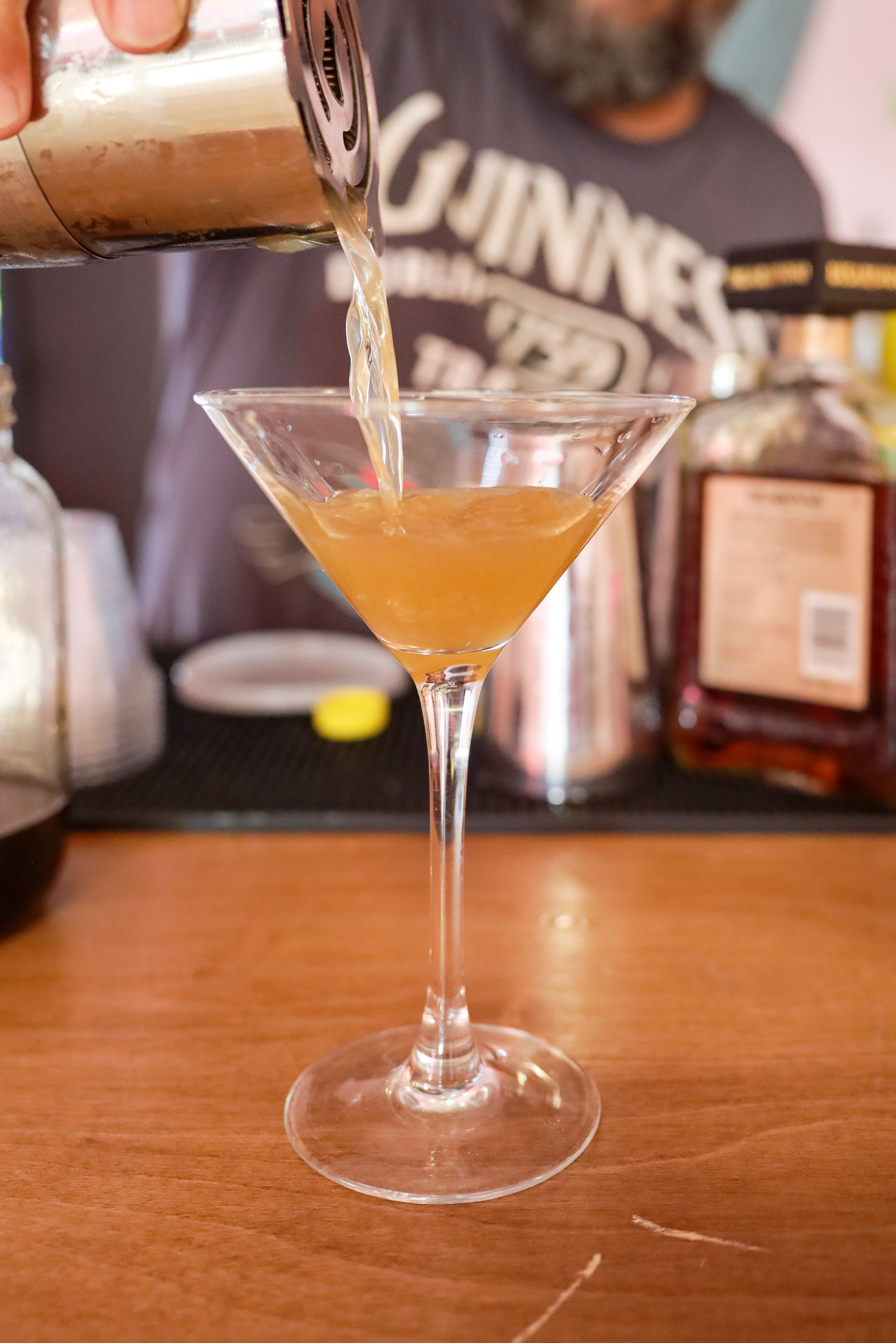 Cocktail being poured into a martini glass on a wooden bar top, with a bartender blurred in the background