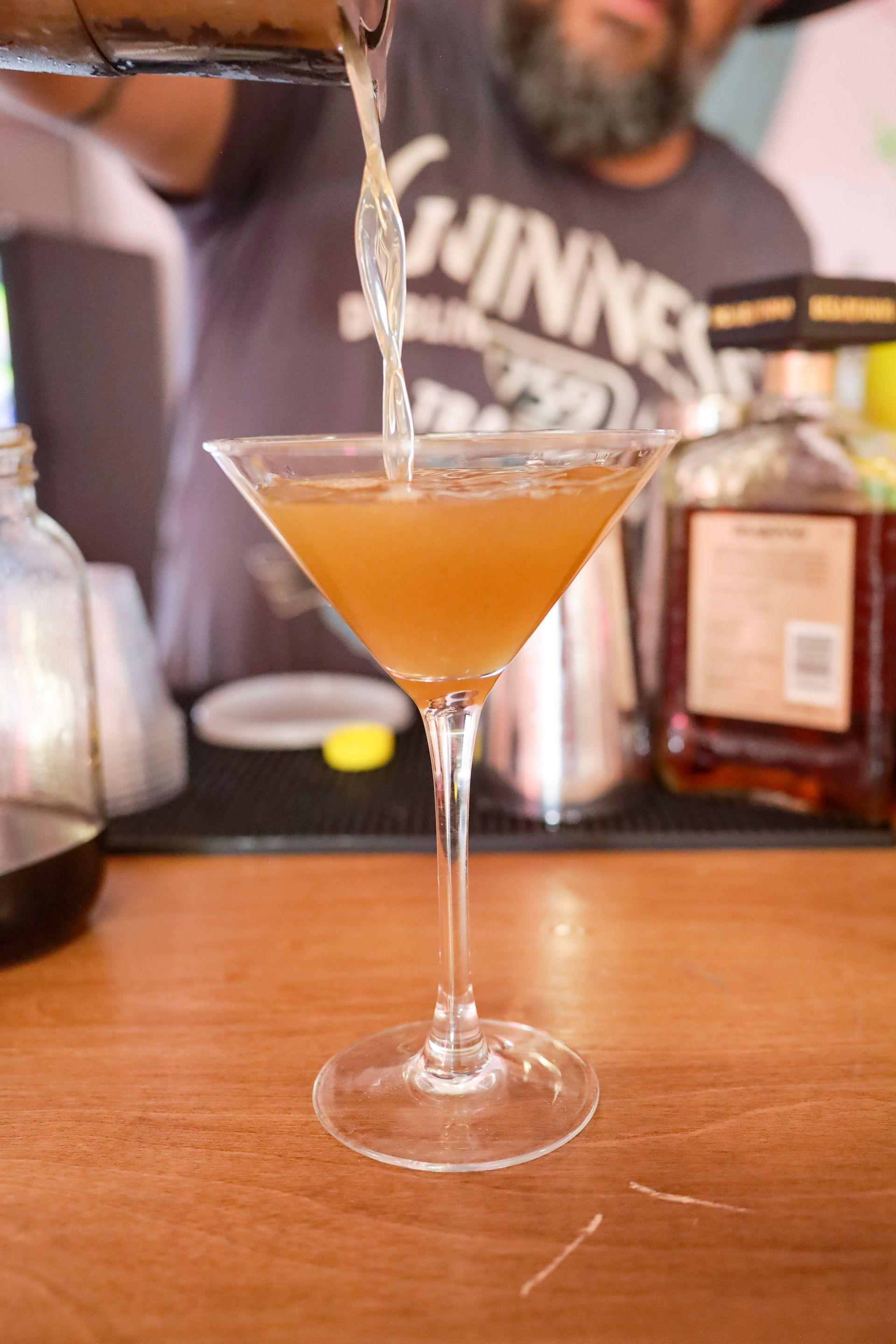 Martini glass with amber cocktail being poured on a bar counter, bottles blurred in the background