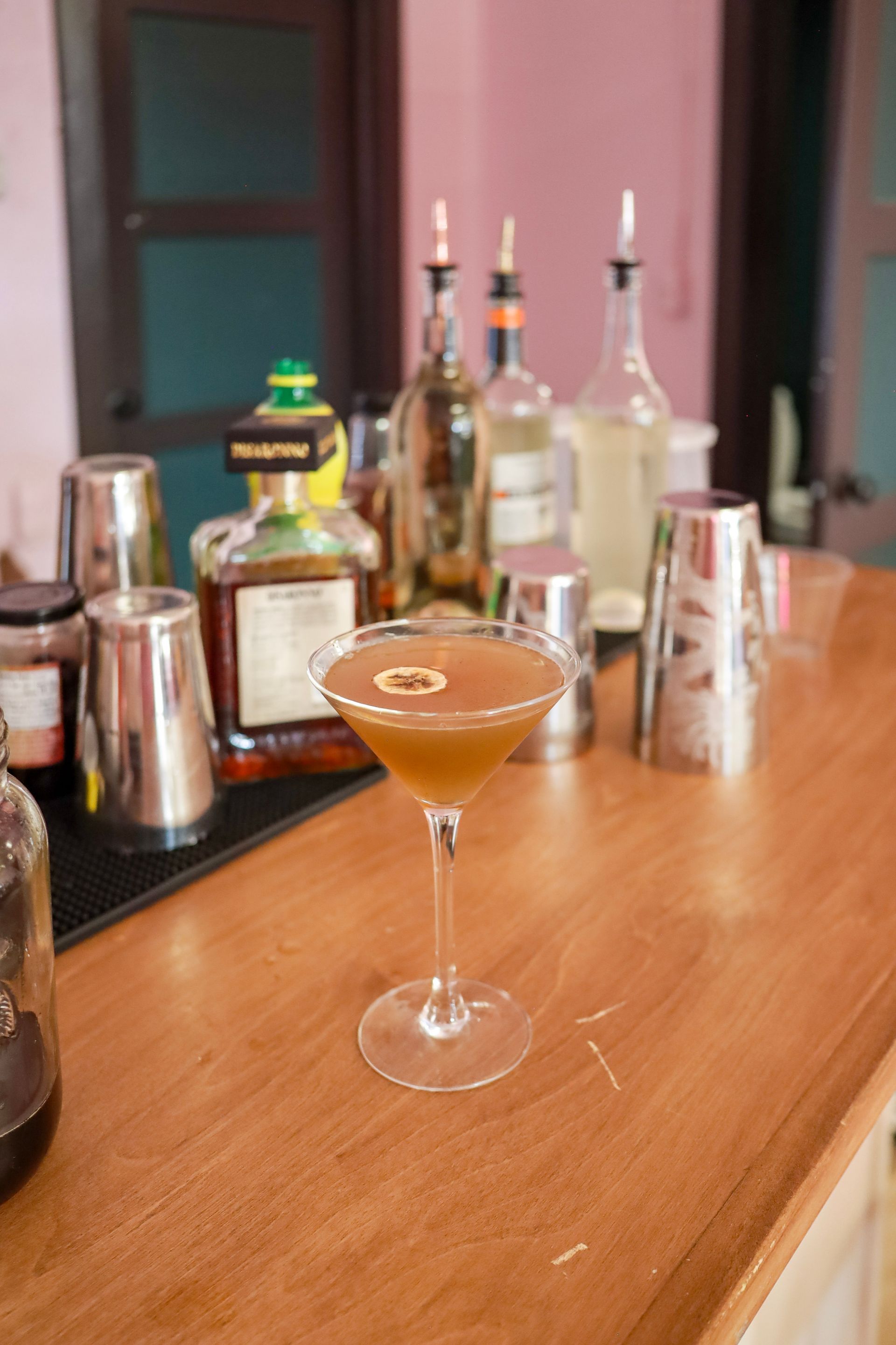 Cocktail in a coupe glass on a bar counter, with bottles and shakers in the background