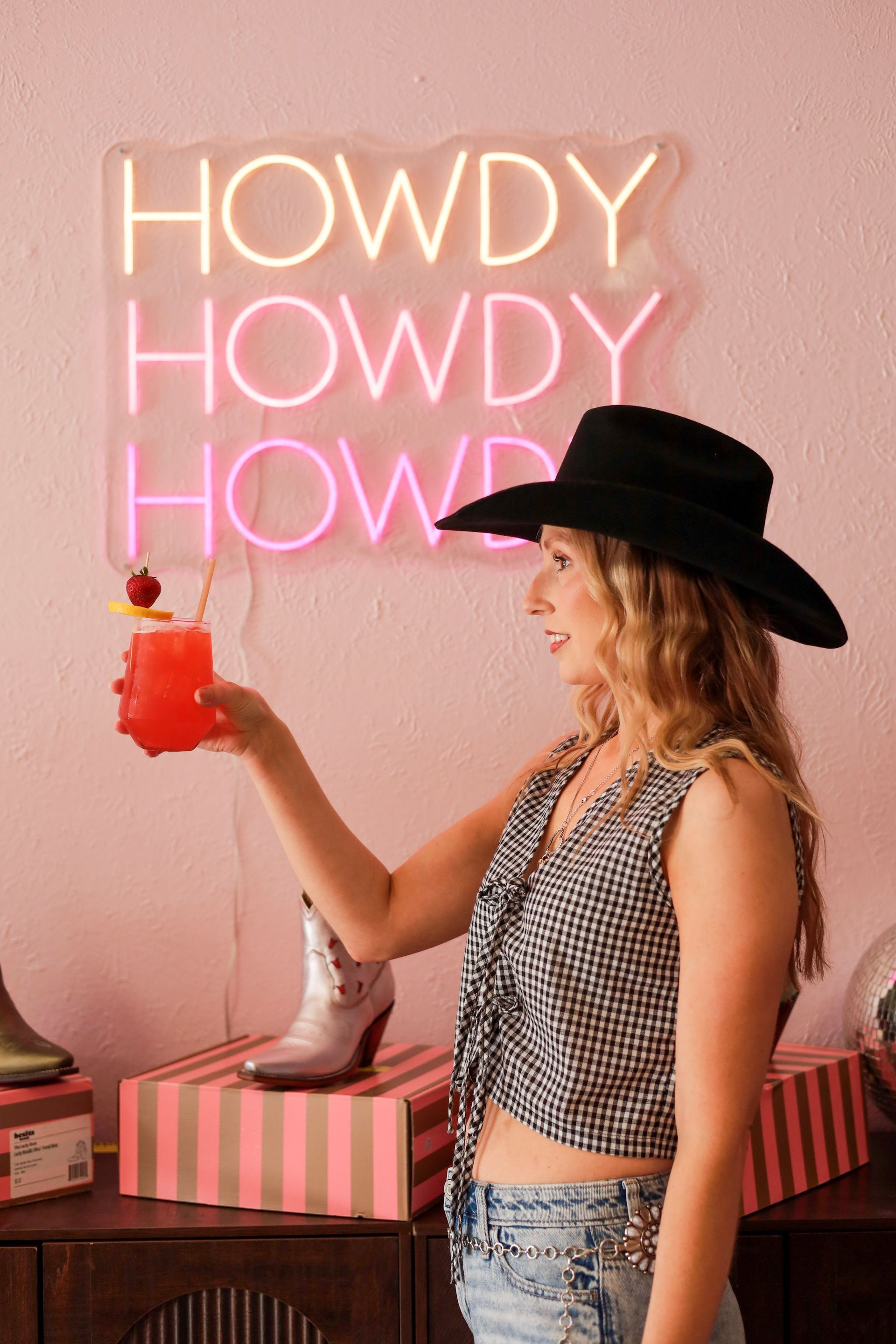 Woman in a black hat holding a red drink beneath pink neon “HOWDY” sign