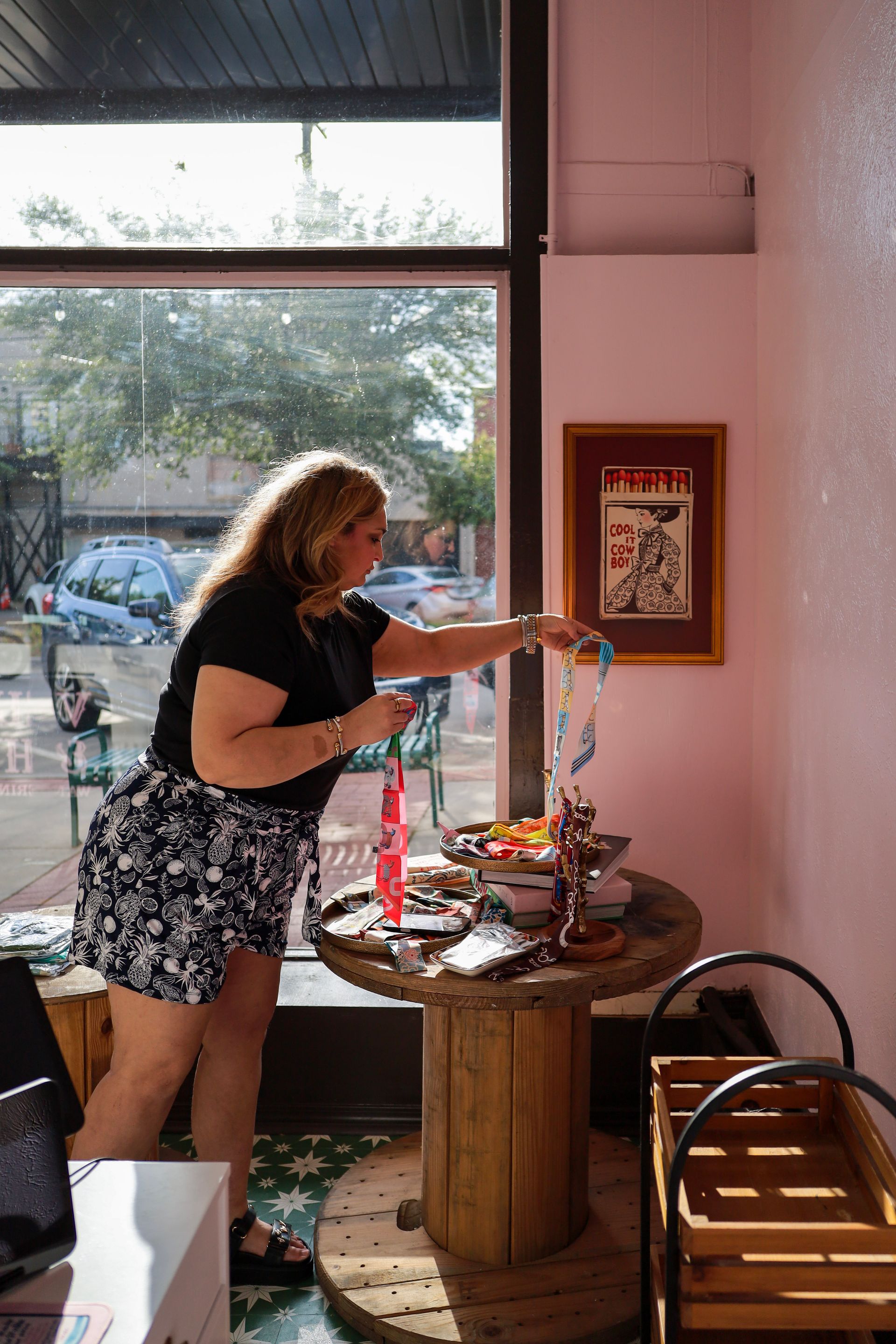 Woman arranging items on a small round table by a large window in a bright cafe or shop