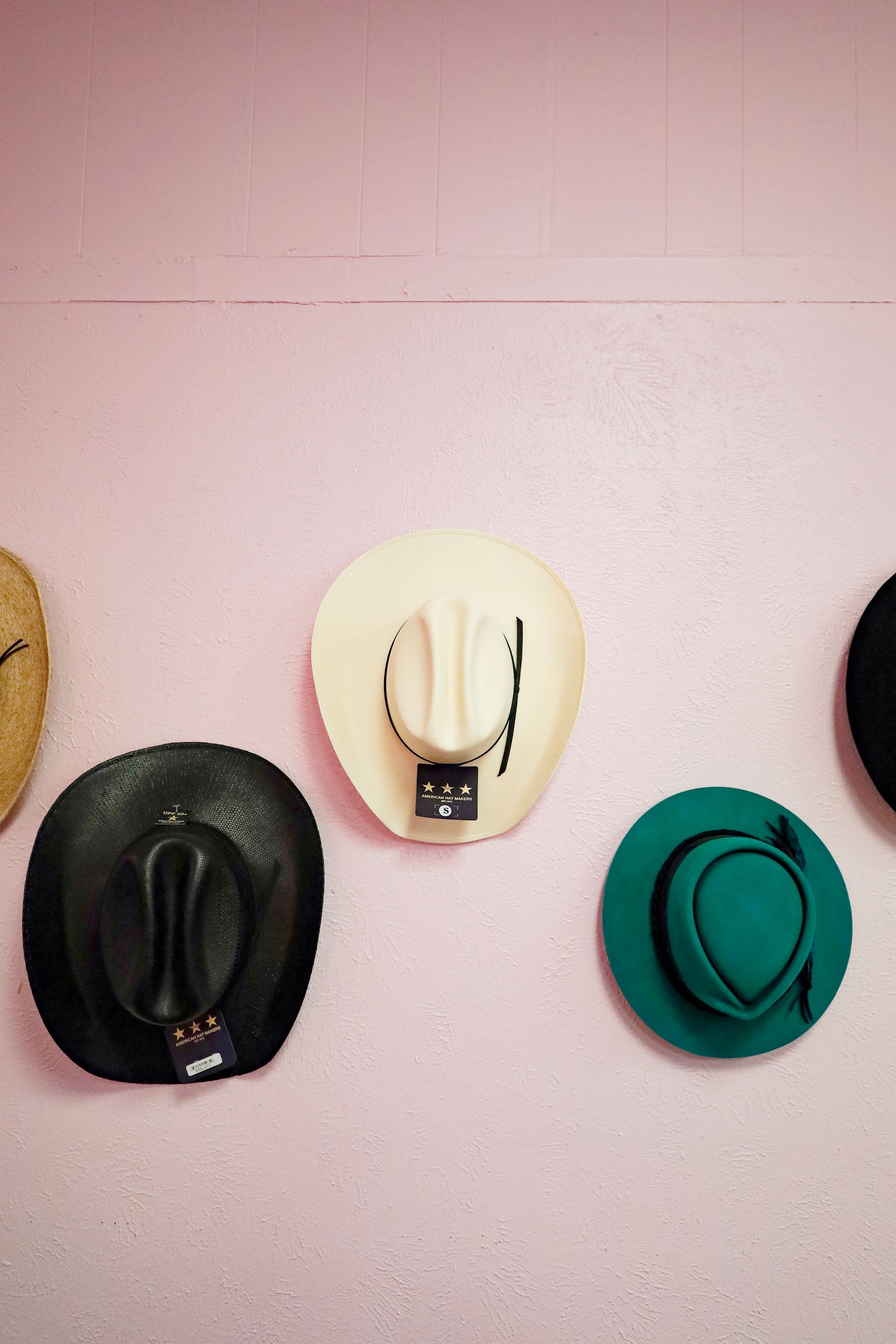 Three hats hanging on a pink wall, including a cream cowboy hat and a green hat.