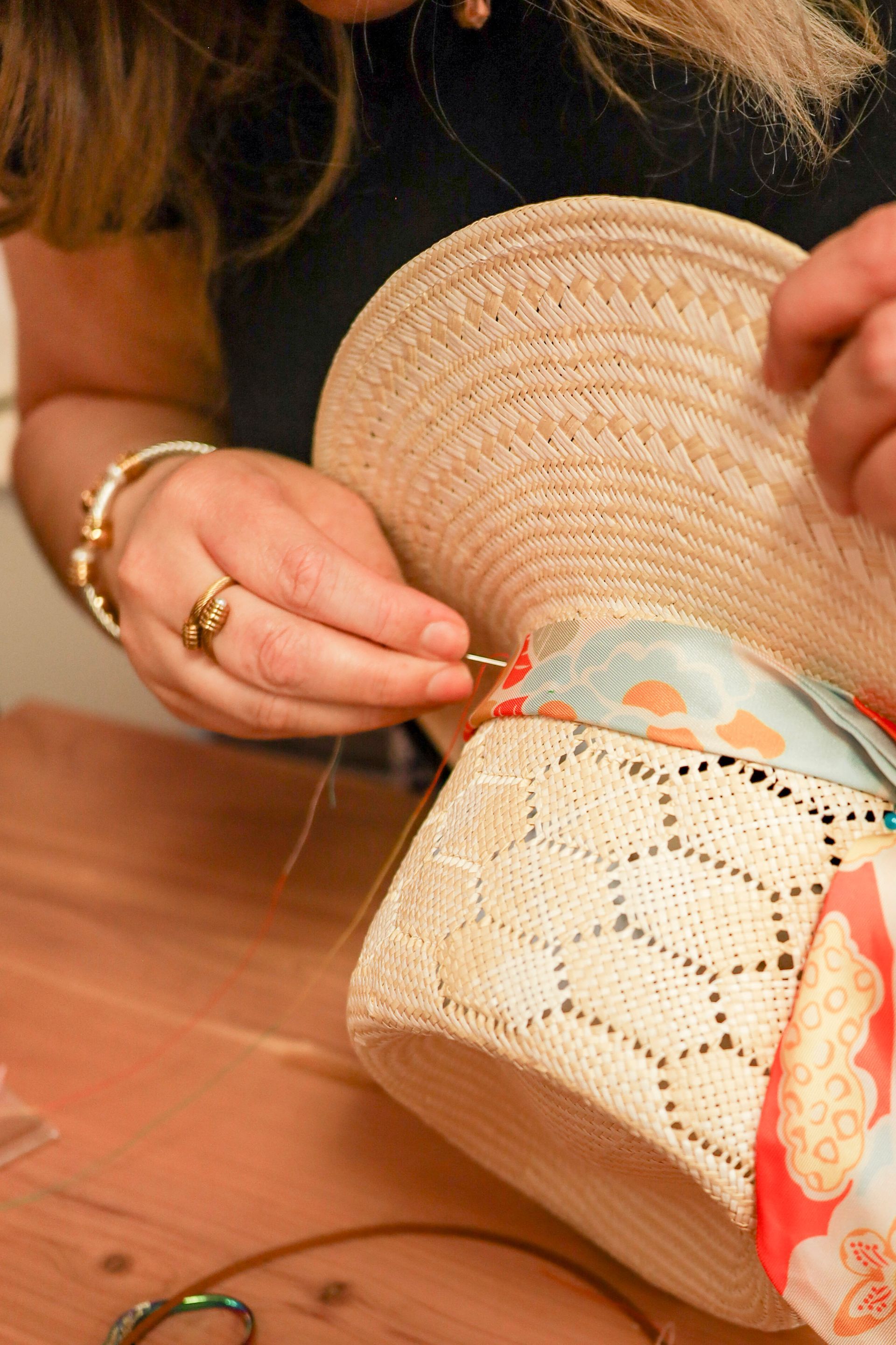 Hands sewing a ribbon onto a straw hat on a wooden table