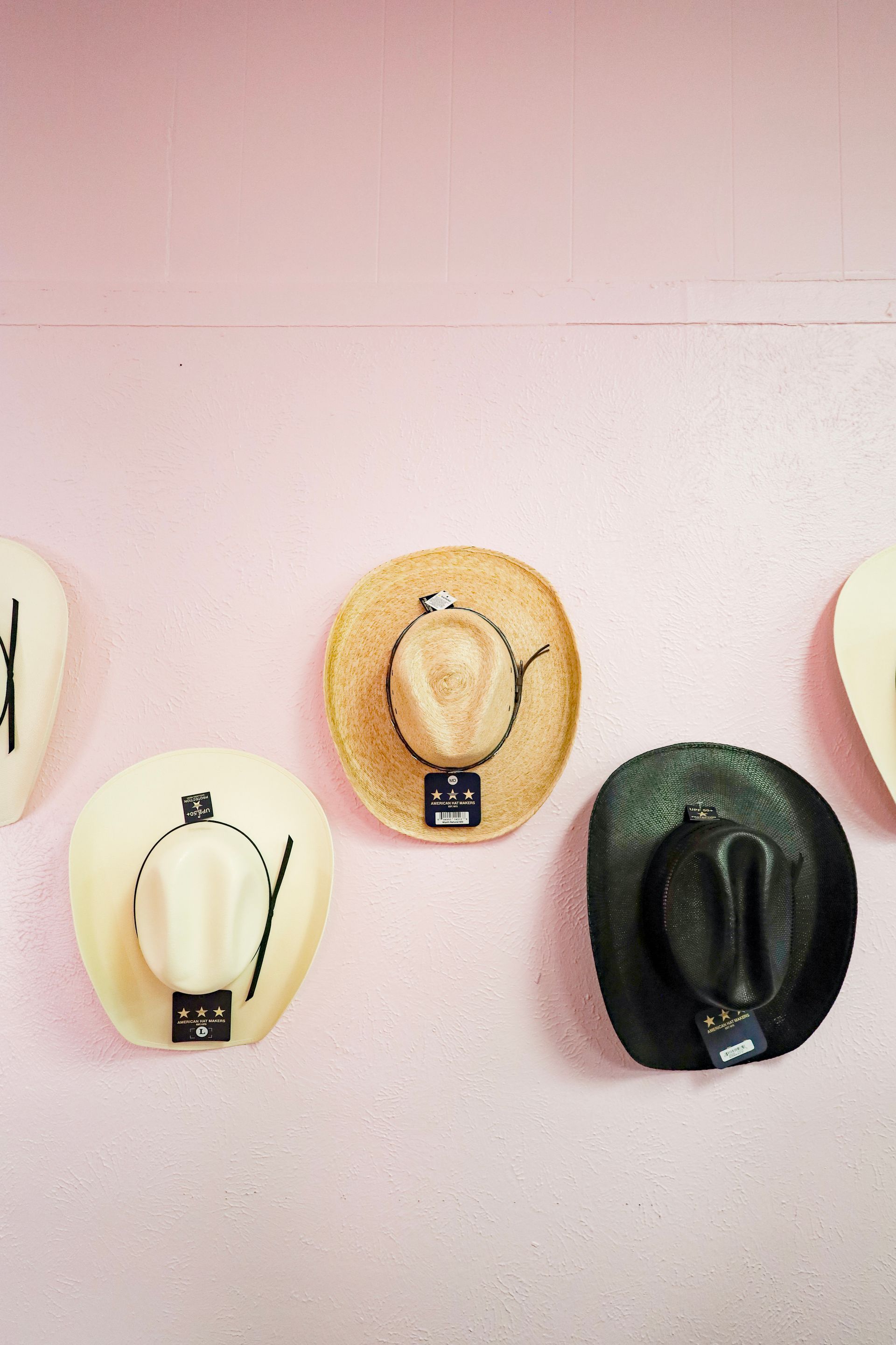 Three hats displayed on a pale pink wall: cream, tan, and black.