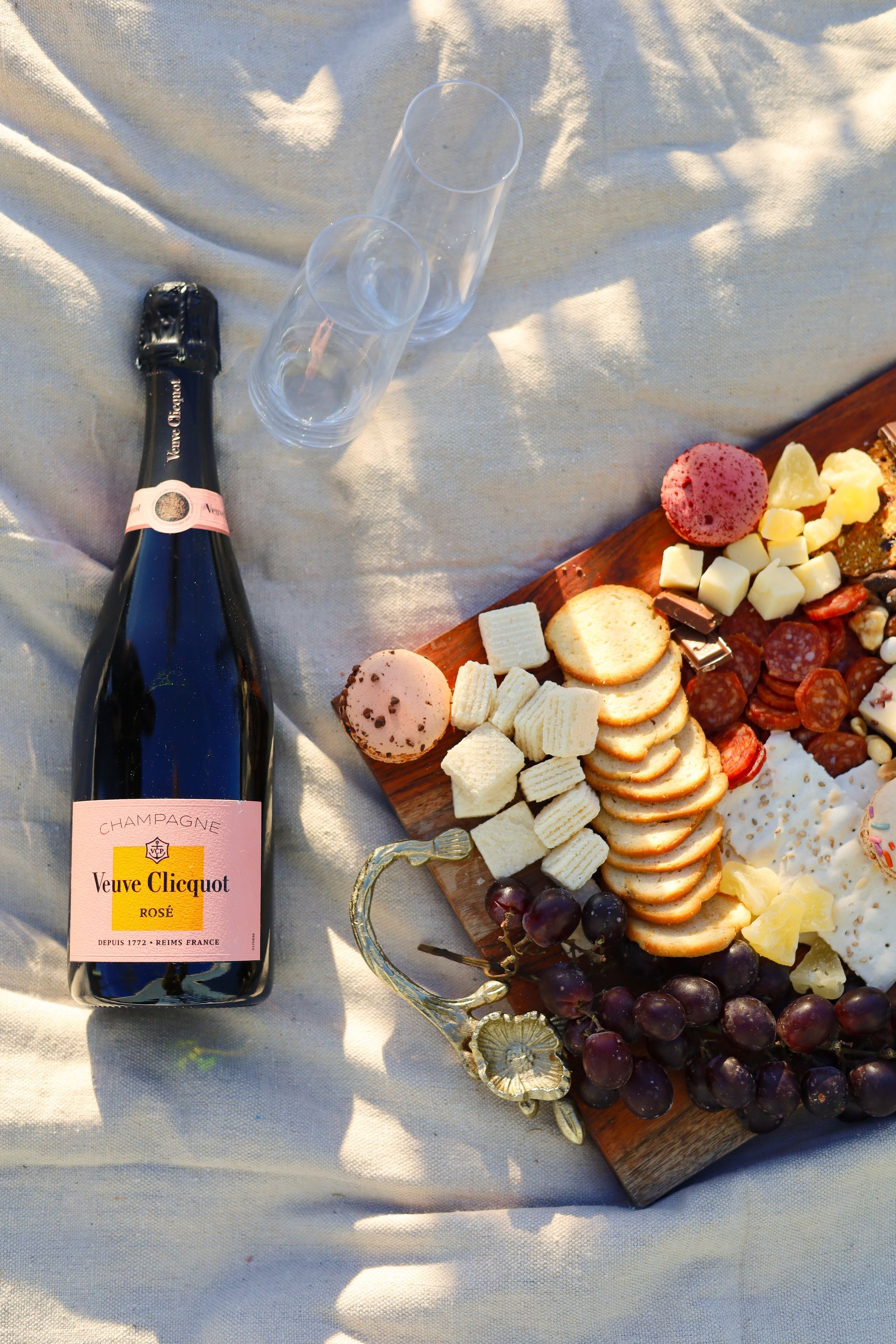 Bottle of sparkling wine beside a charcuterie board with fruit, cheese, crackers, and cured meats on a blanket