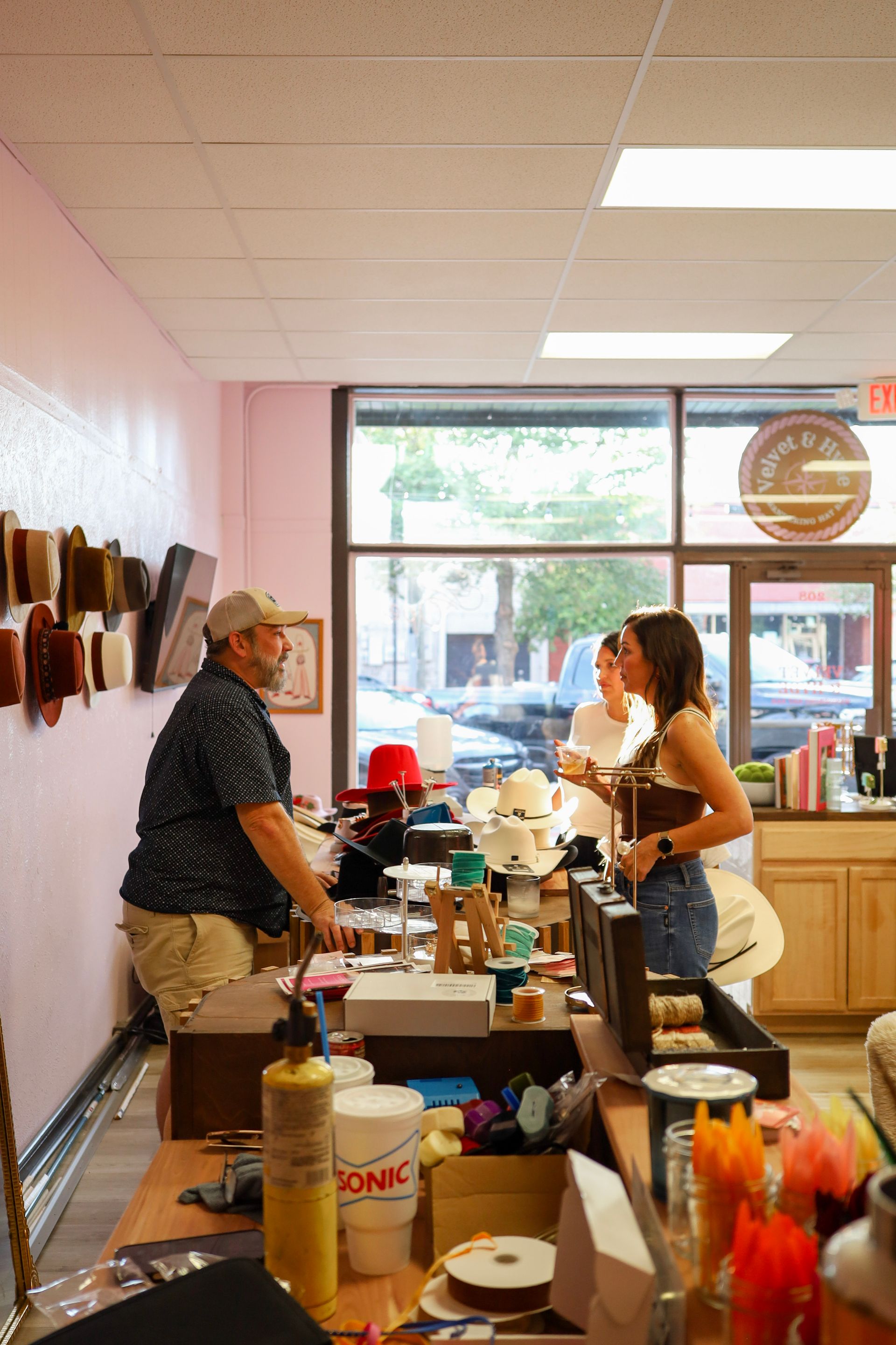 Two people chatting across a counter in a cozy café with bright windows and pastries on display