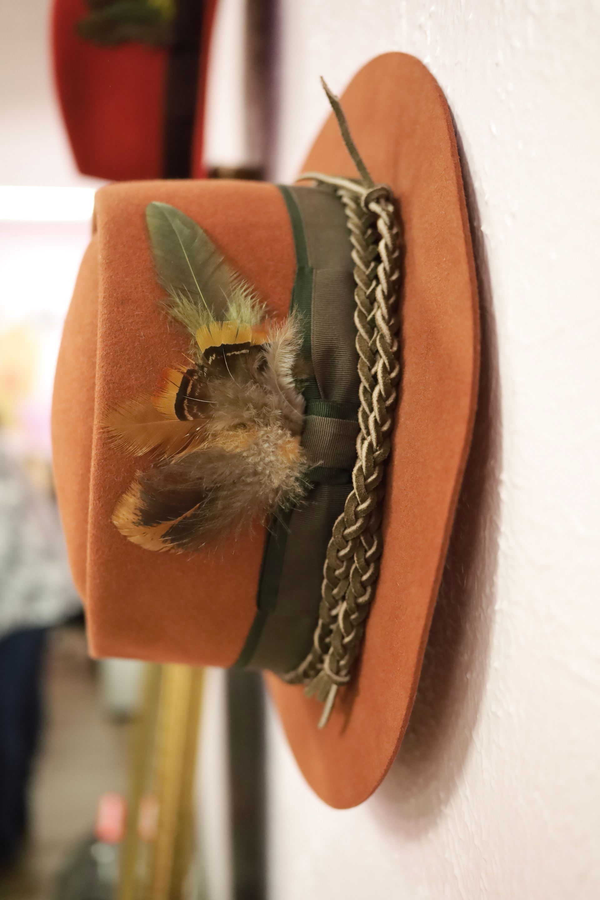 Brown hat with a feather and braided trim hanging on a wall hook