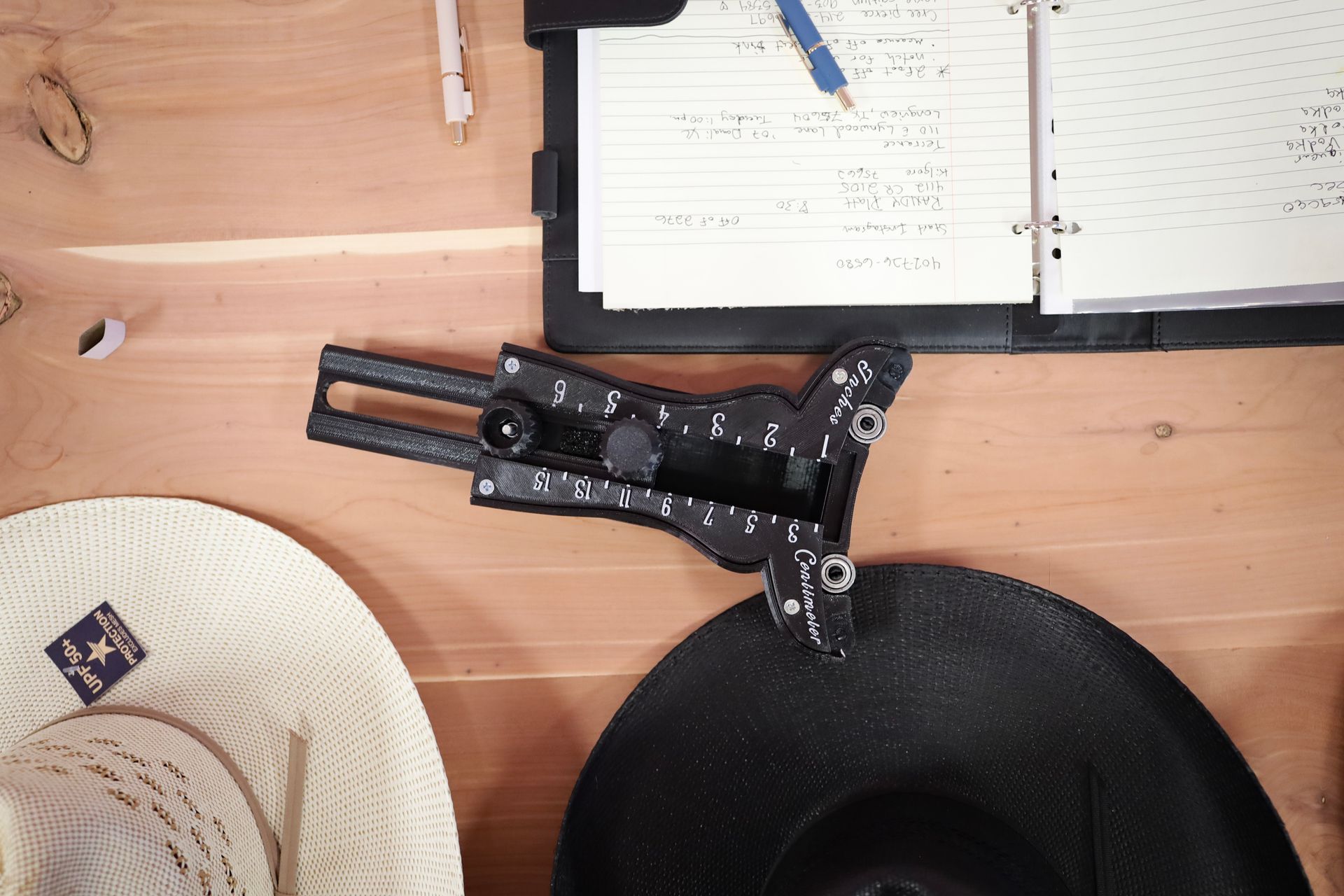 Overhead view of a wooden desk with a notebook, pens, and a black mechanical arm over a hat