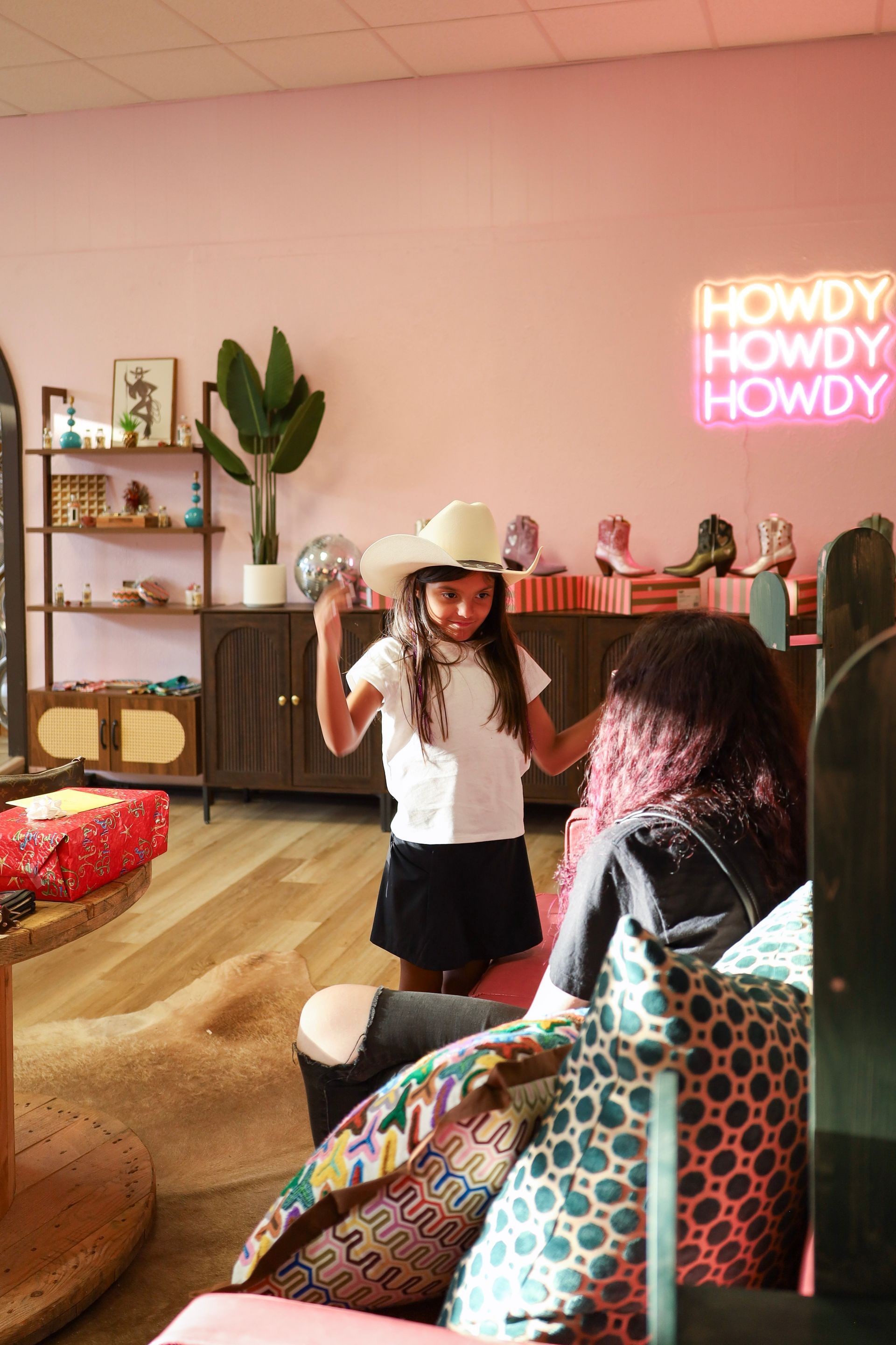 Woman in white hat speaking in a pink room with neon “HOWDY” sign and shelves in the background