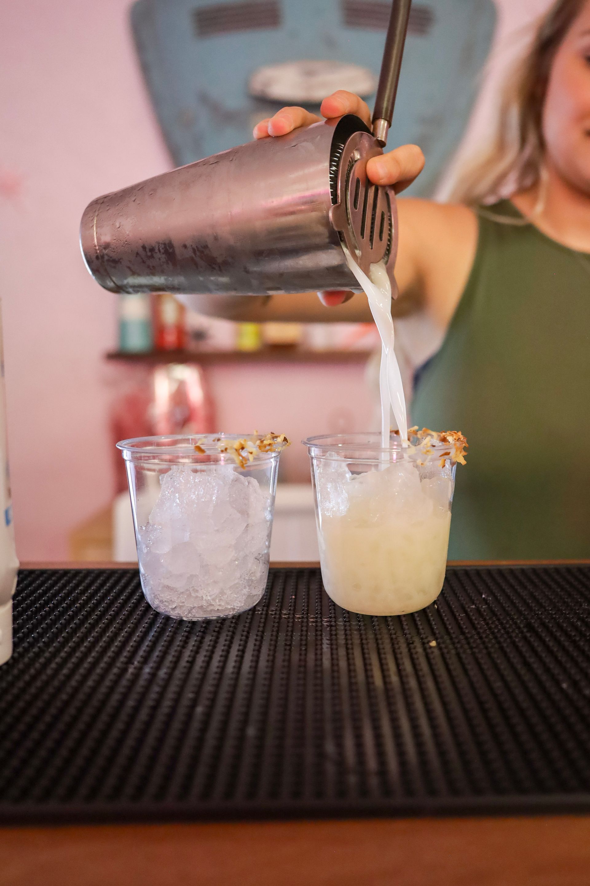 Bartender pouring cocktails from a shaker into two clear glasses on a bar mat