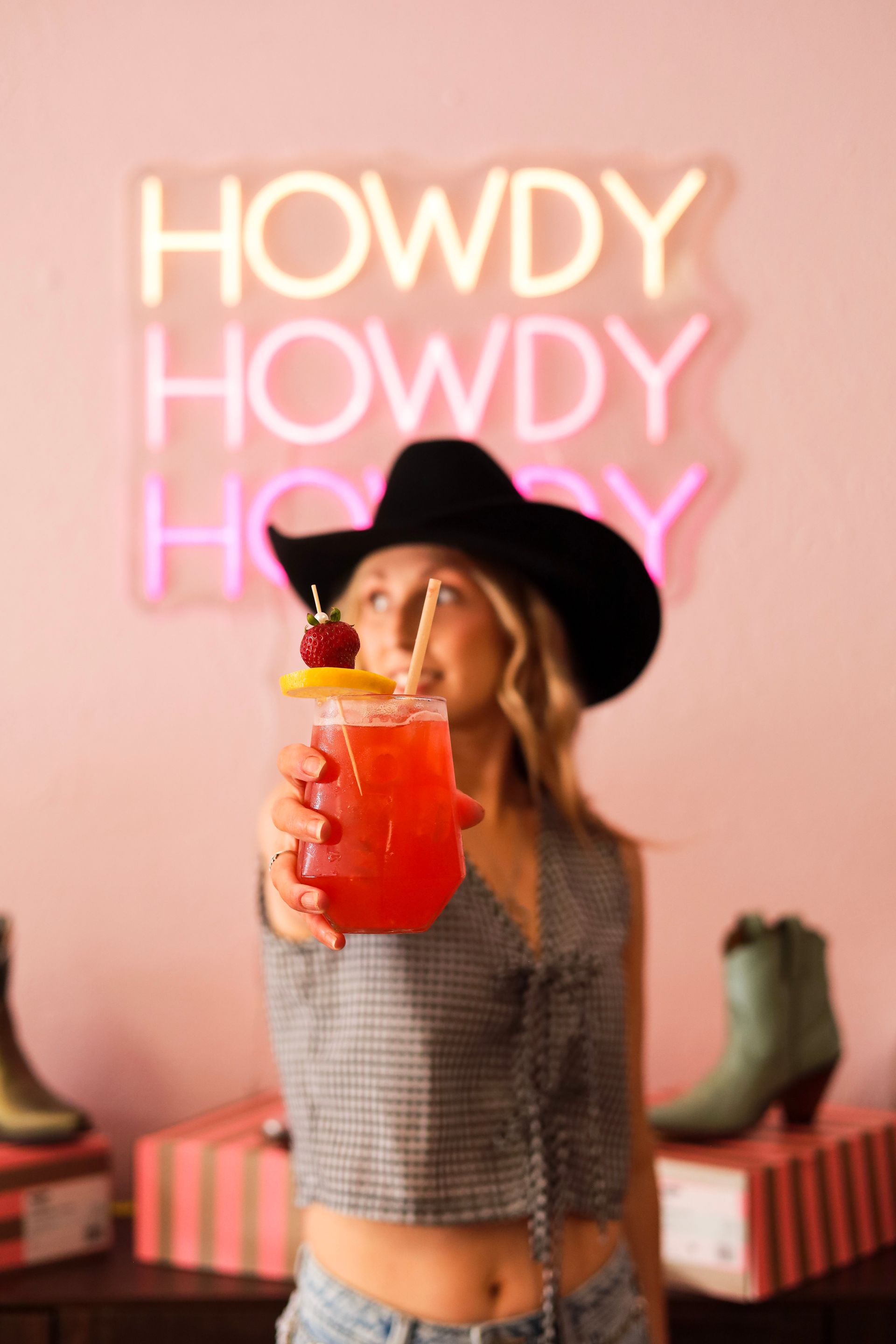 Woman in black hat holding a red drink in front of a neon “HOWDY” sign