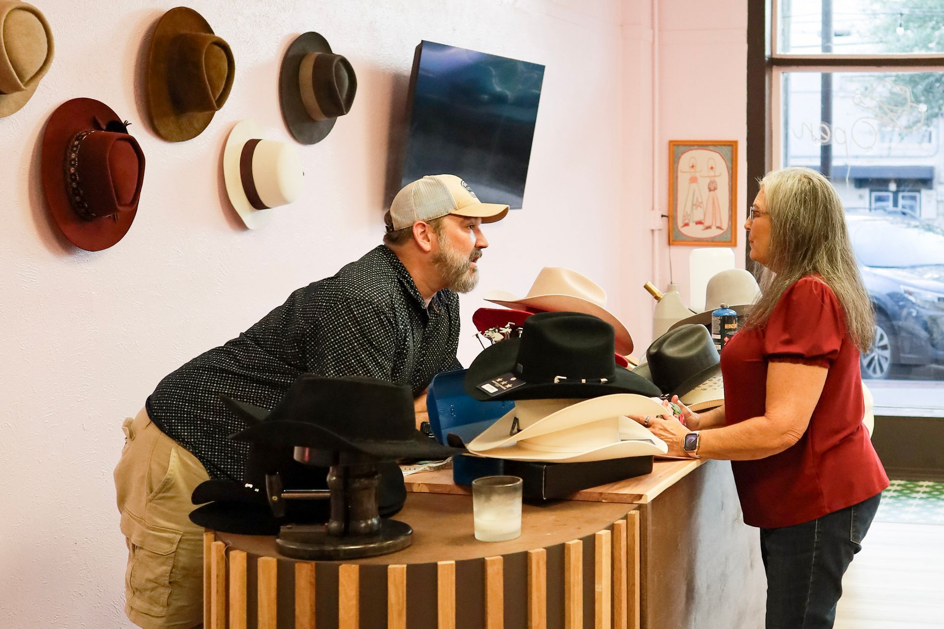 Two people talking across a counter in a hat shop, with several hats displayed on the wall.