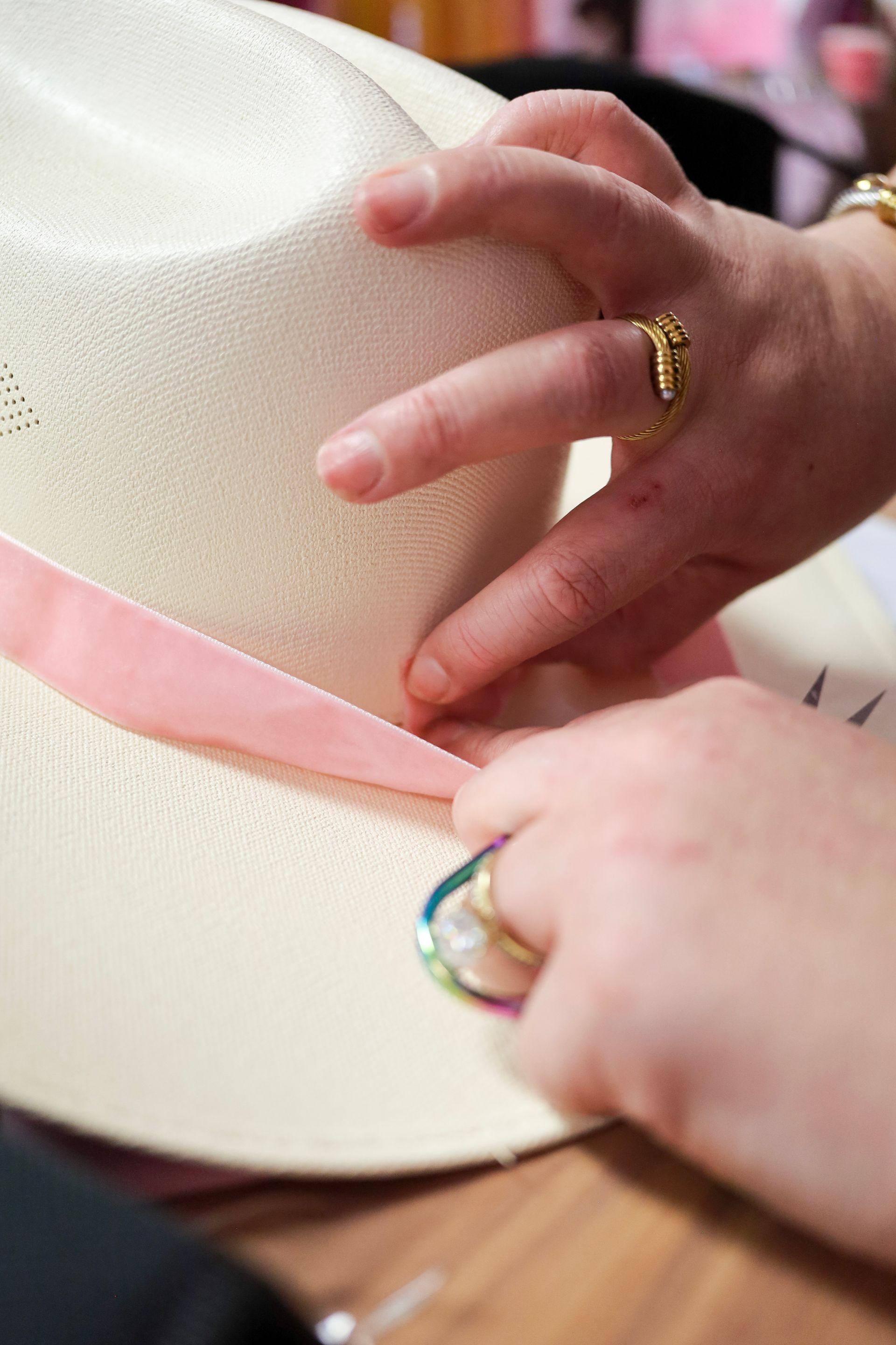 Hands placing a pink ribbon around a cream-colored hat on a table
