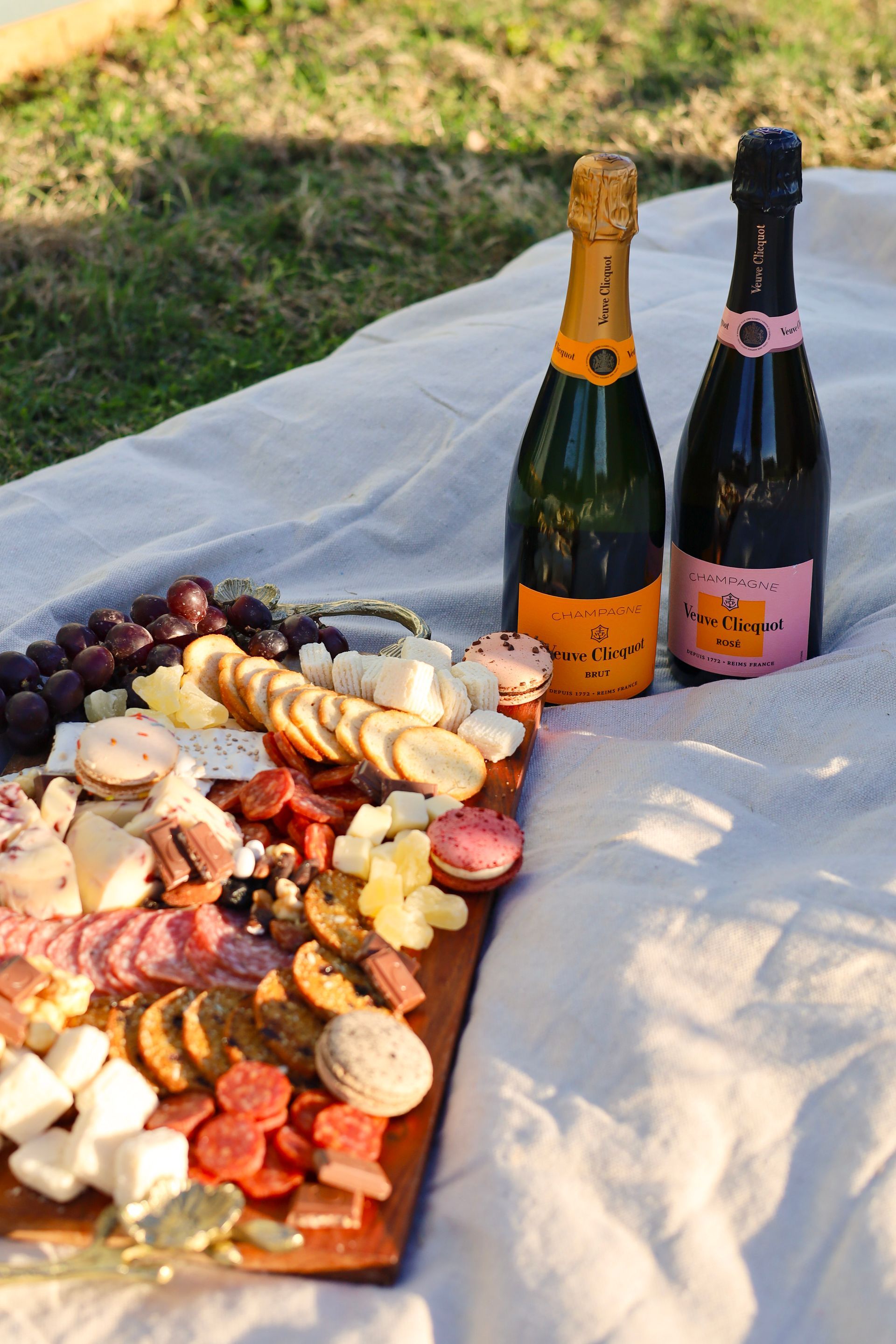 Charcuterie board with crackers and fruit beside two bottles of sparkling wine on a picnic blanket