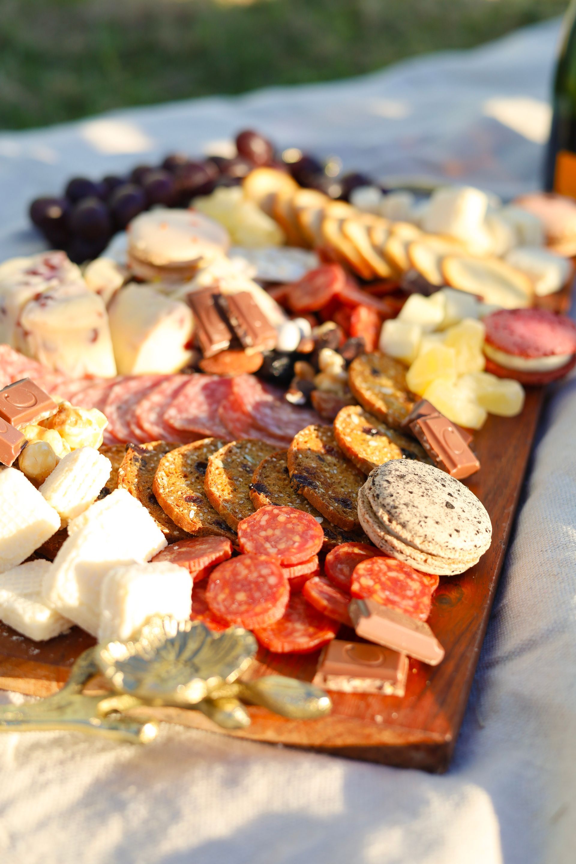 Assorted cheeses and crackers on a wooden serving board outdoors, with grapes and sliced meats