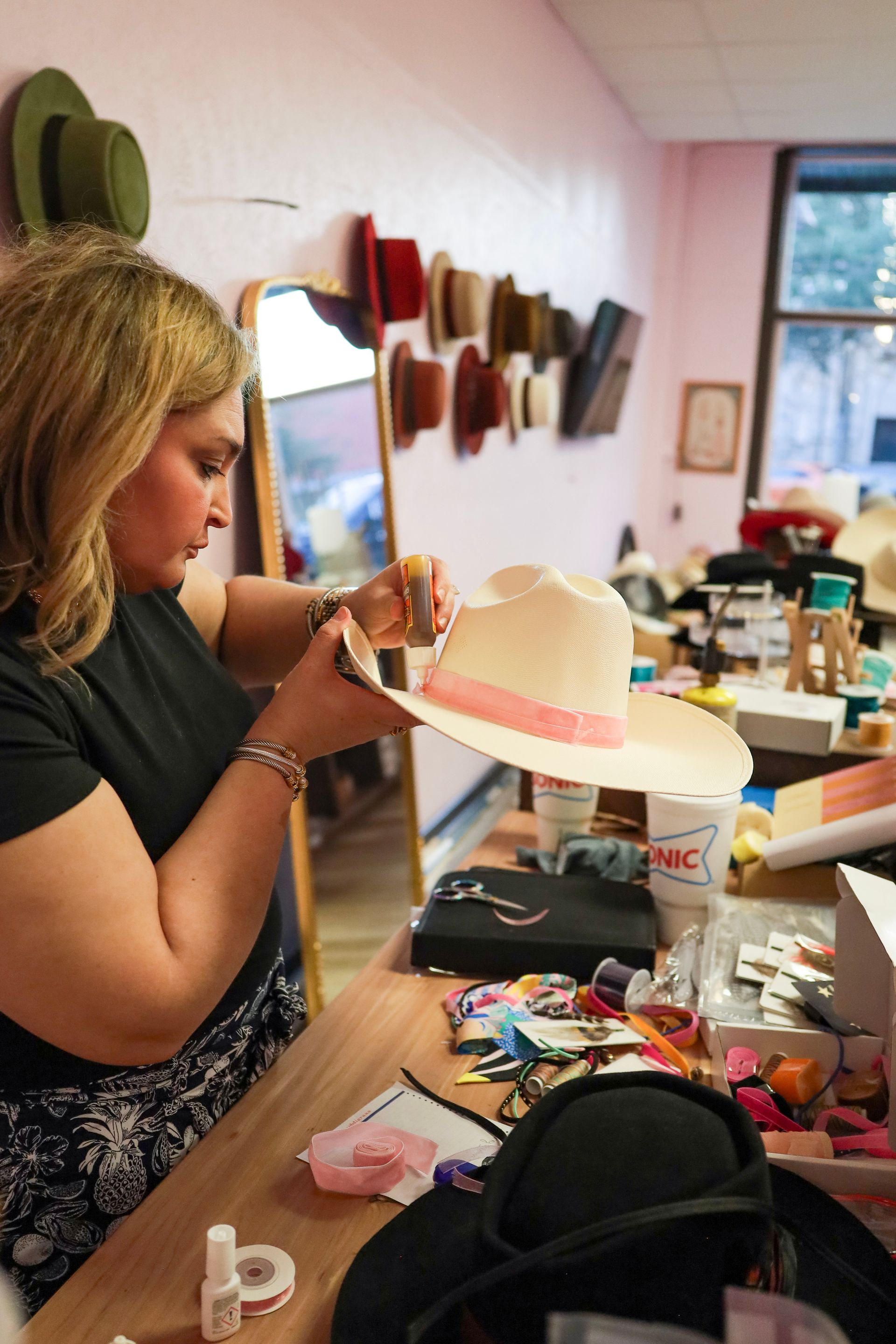Person painting a pink hat at a cluttered craft table in a workshop studio