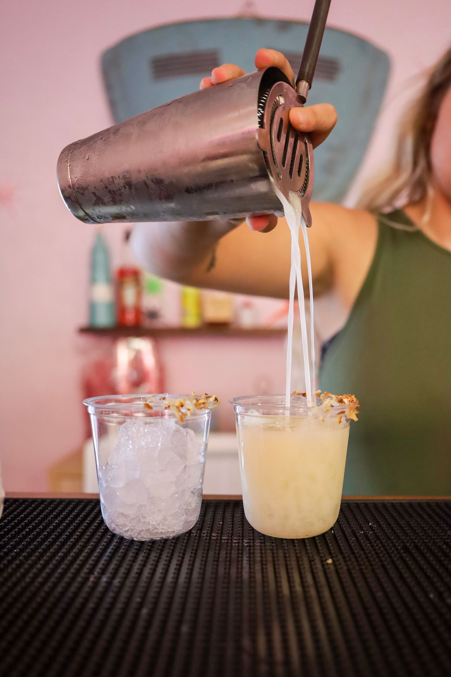 Bartender pours a pale drink from a shaker into a glass beside an ice-filled glass on a bar mat