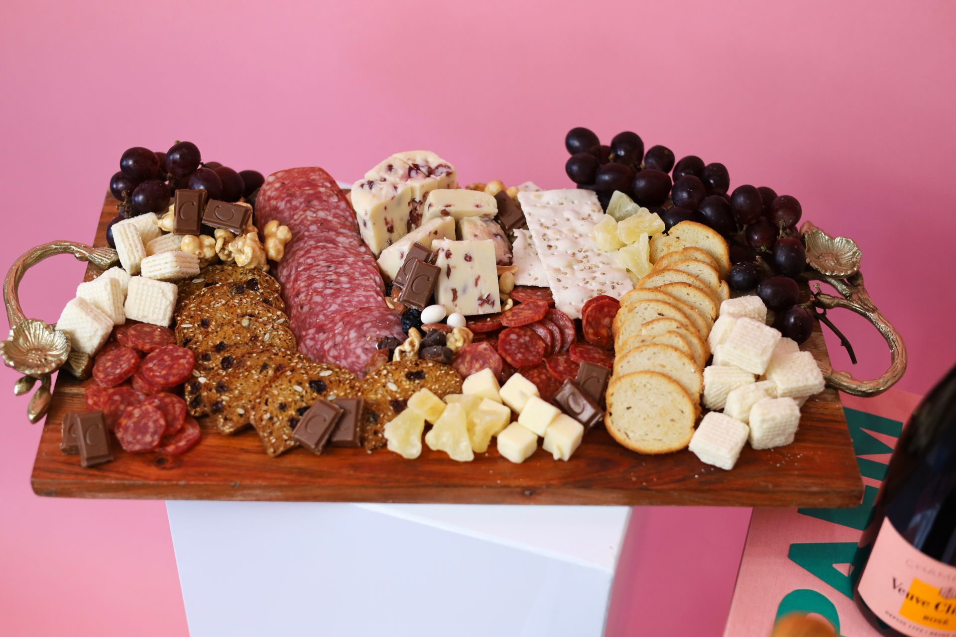 Charcuterie board with crackers, cheeses, meats, grapes, and sweets on a wooden tray against a pink background