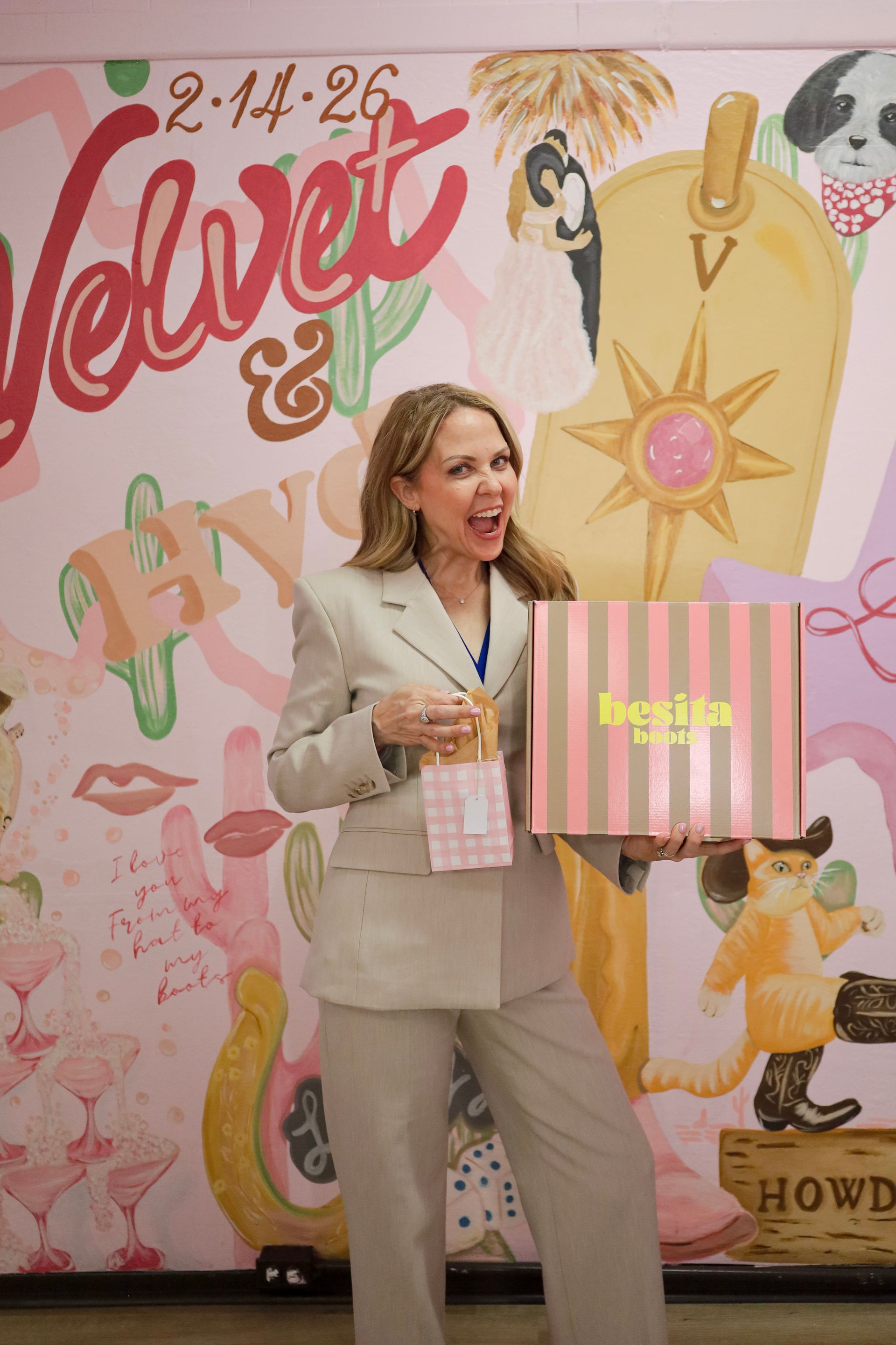 Woman holding a pink striped shopping bag in front of a colorful mural and wearing beige clothing