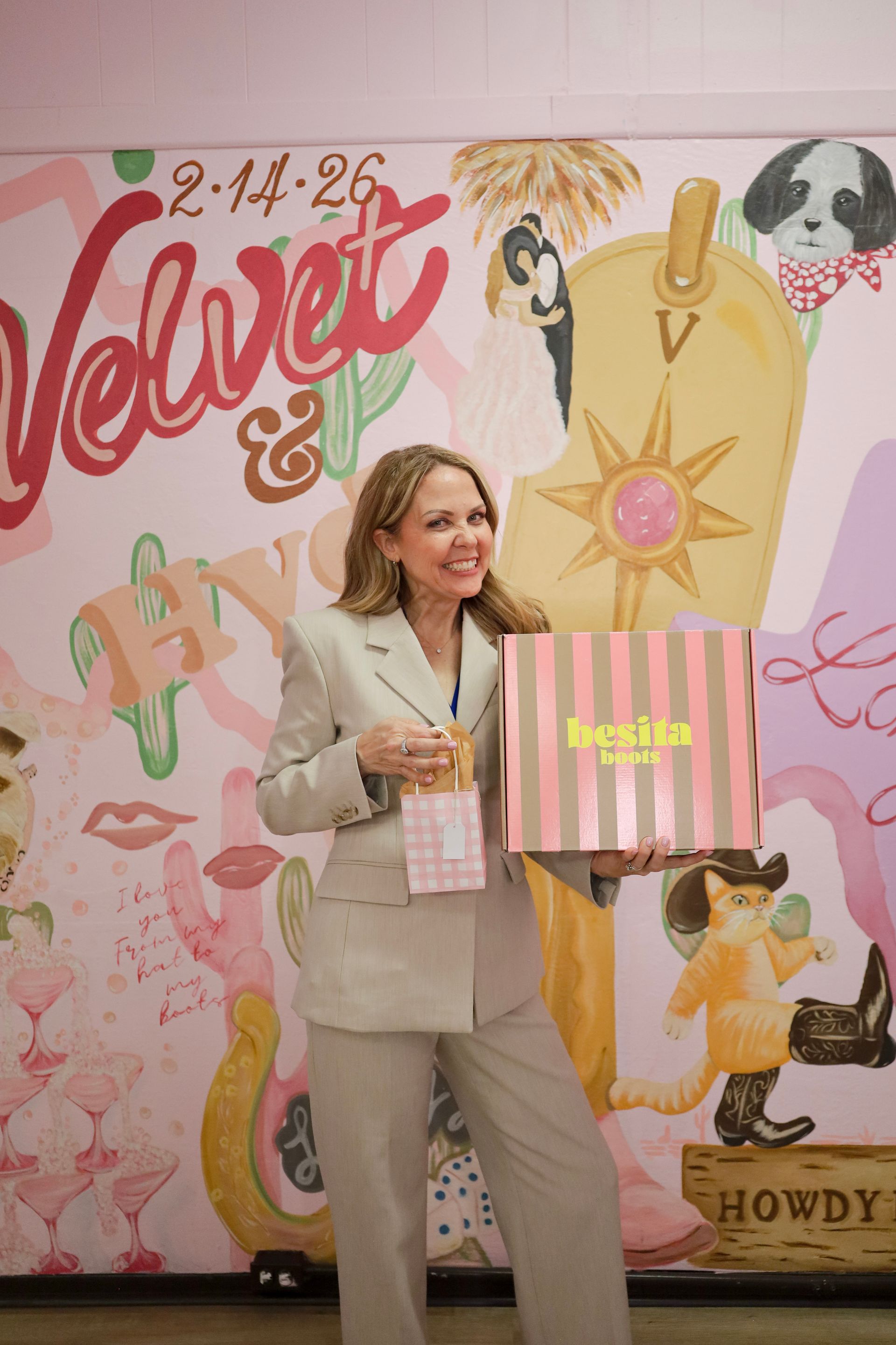 Woman in beige suit posing with a shopping bag in front of a colorful Velvet & Bow backdrop.