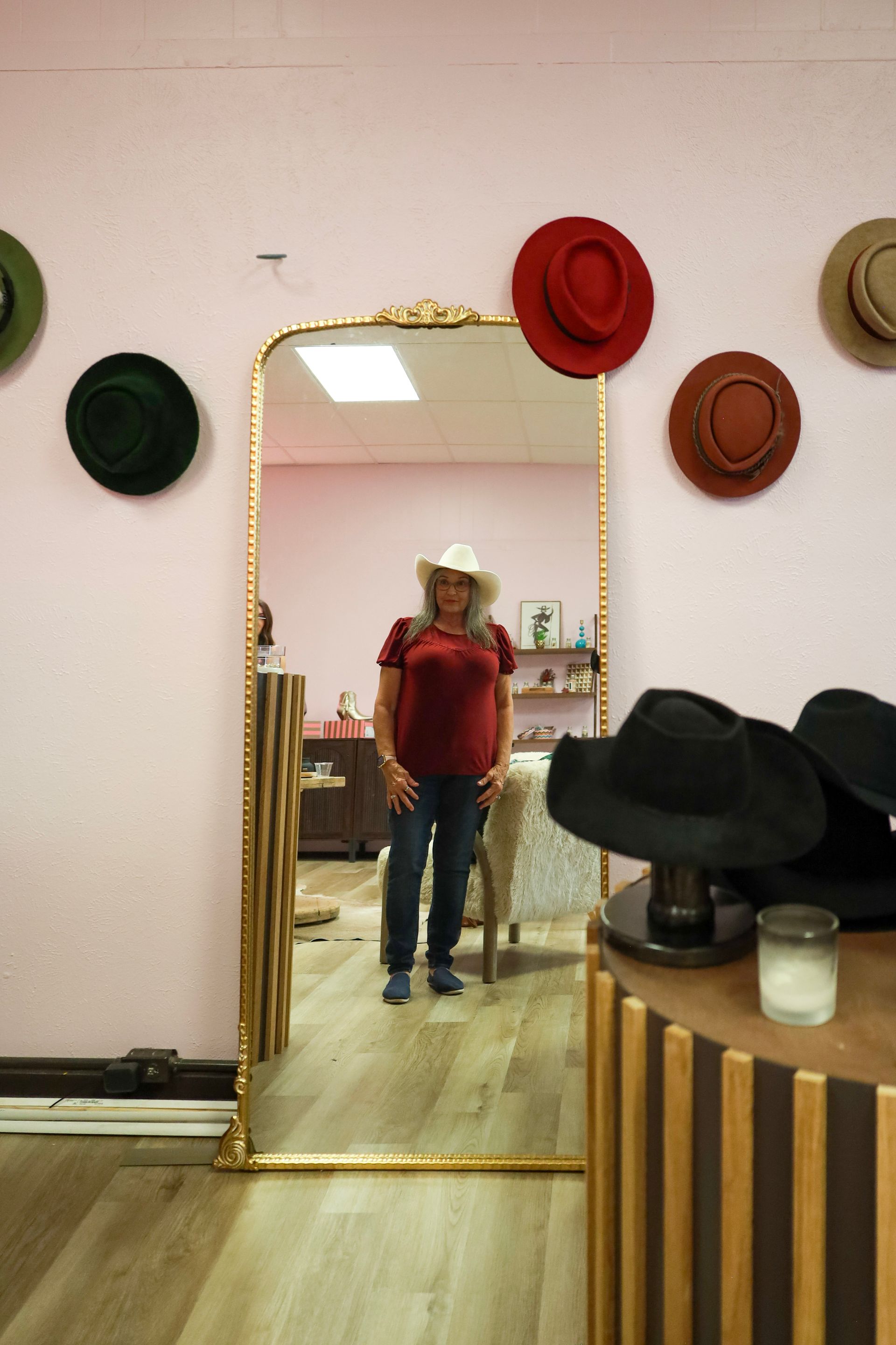 Person in a red shirt and hat standing before a gold-framed mirror in a hat shop