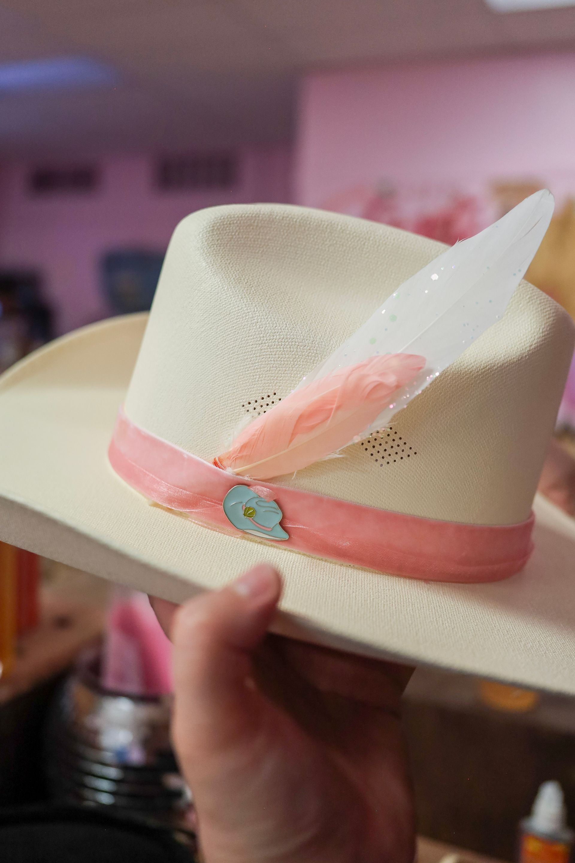 Cream cowboy hat with pink ribbon, feather, and flower detail being held indoors