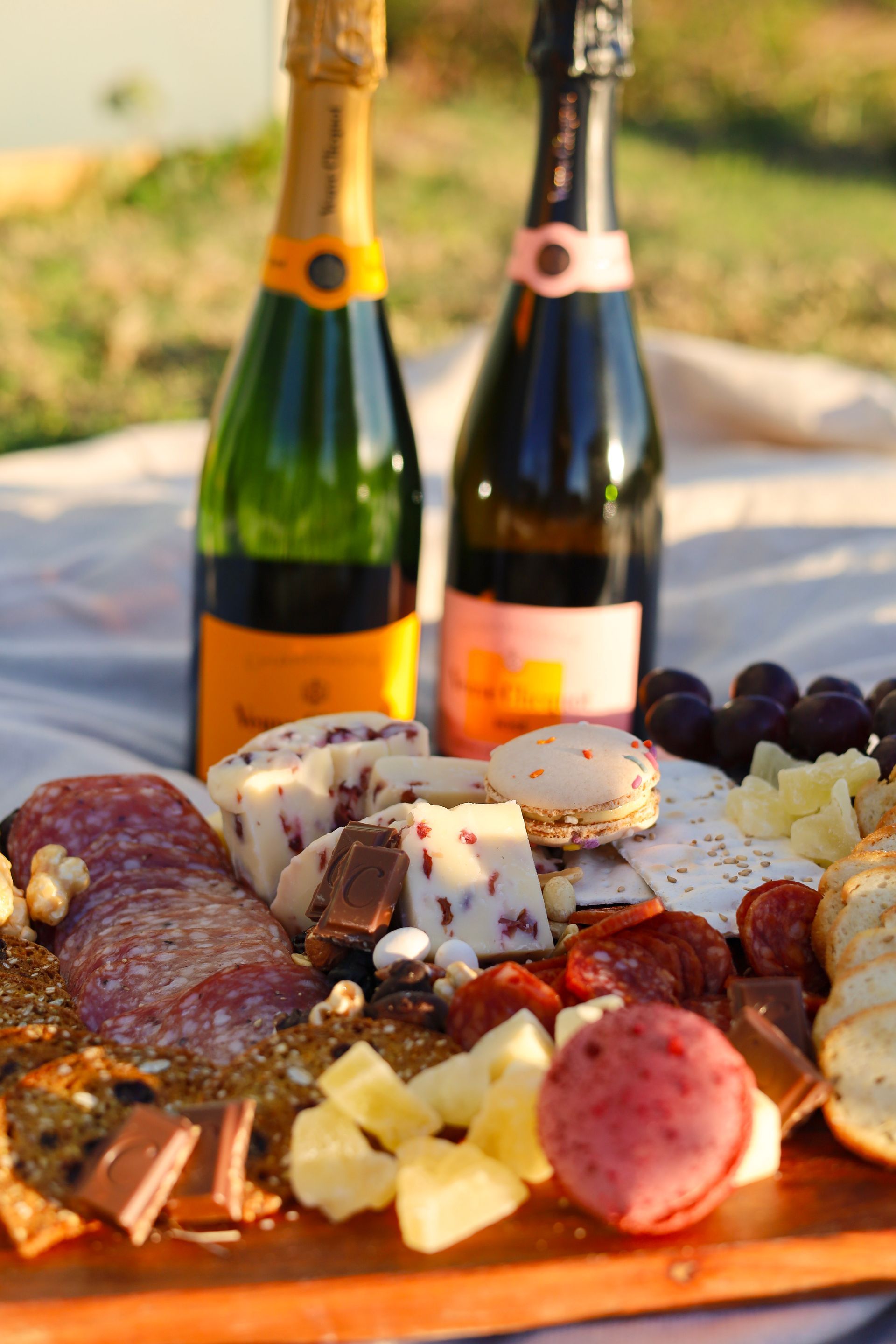 Cheese and charcuterie board with grapes and crackers, with two champagne bottles in the background outdoors