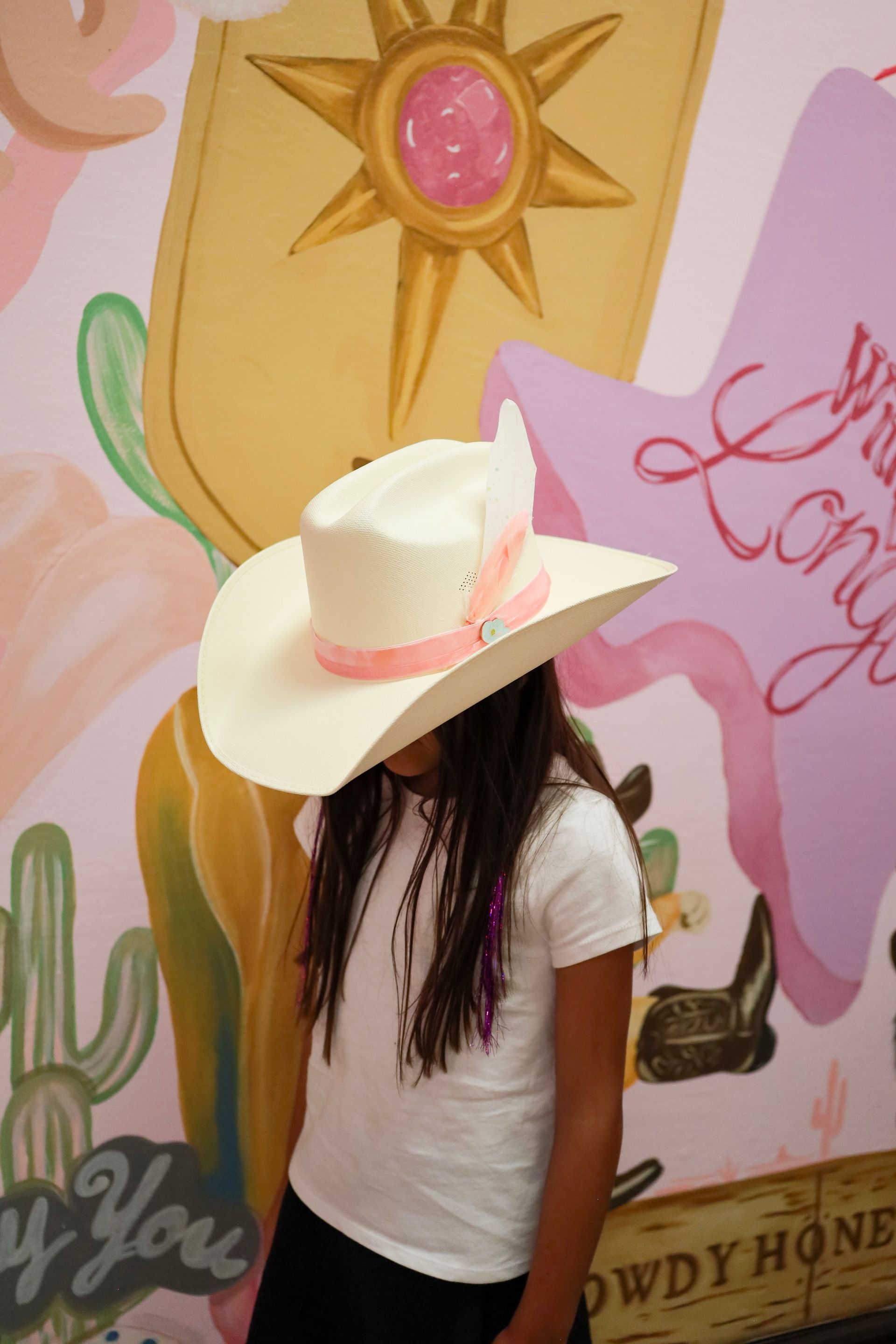 Person in a white hat and shirt poses before a colorful mural with a sun and pink wings.