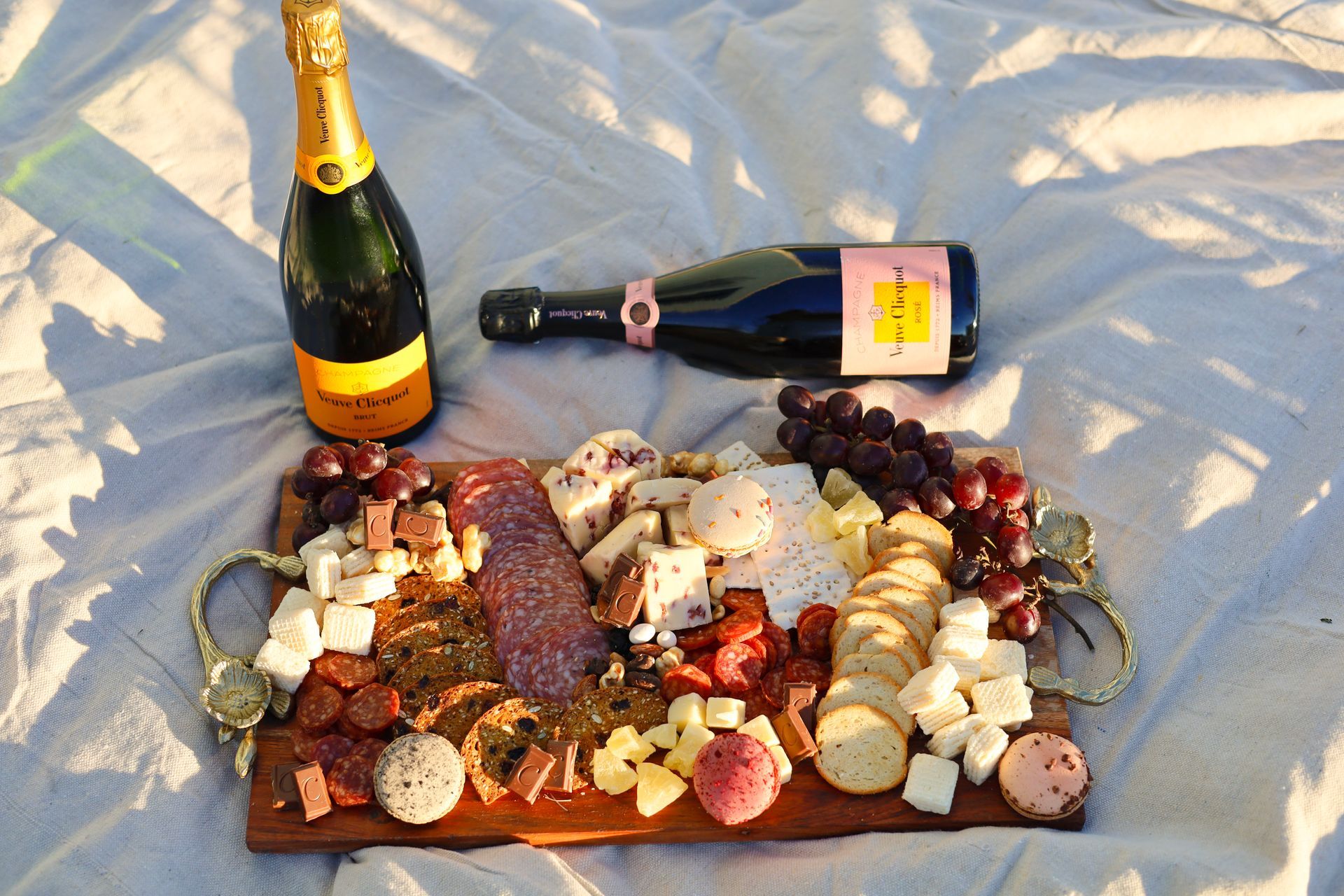 Wine bottles beside a charcuterie spread on a blanket outdoors