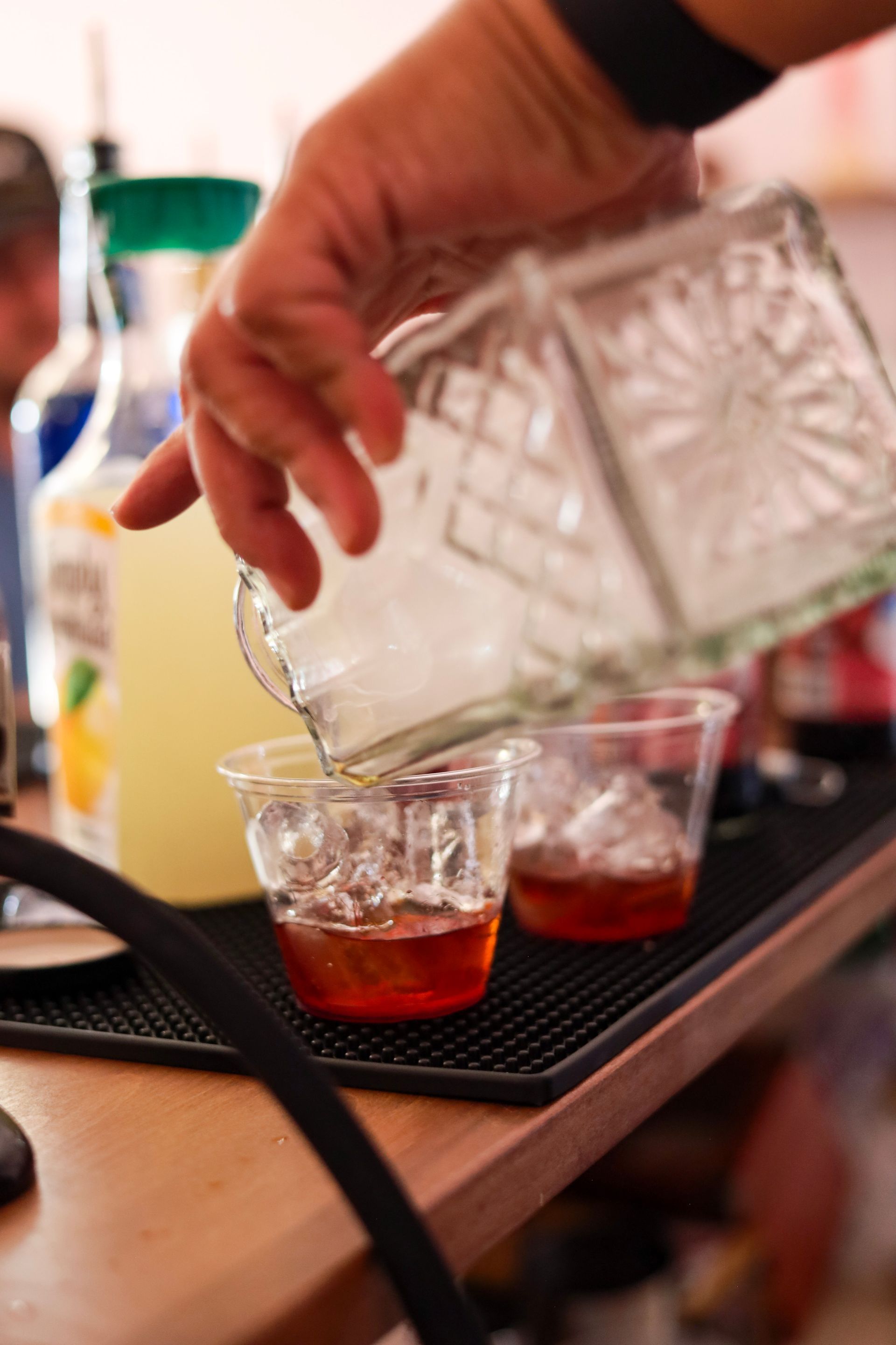 Hand pouring a clear drink from a glass pitcher into a whiskey glass with ice and red liquid on a tray