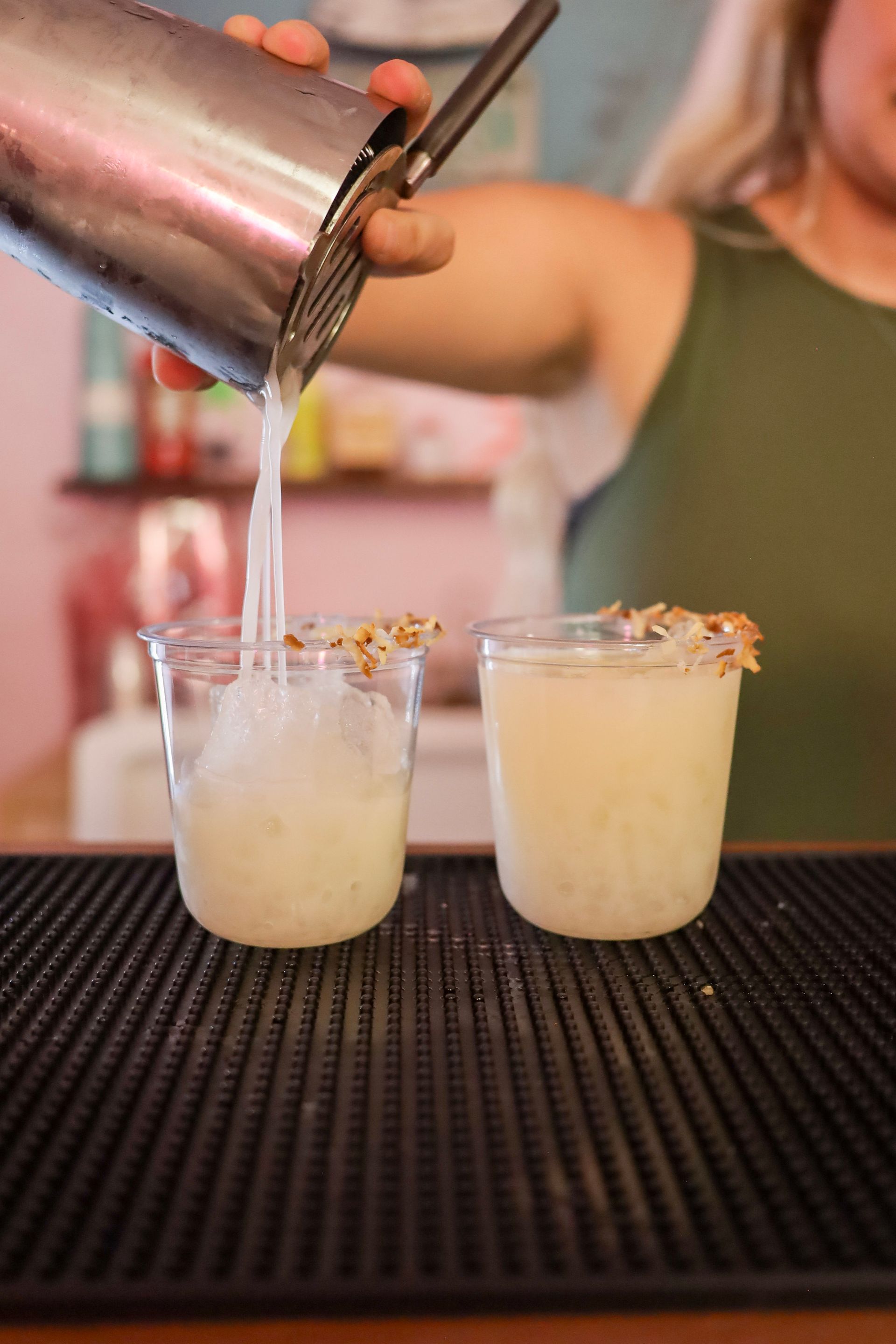 Bartender pouring a creamy cocktail from a shaker into two rimmed glasses at a bar.