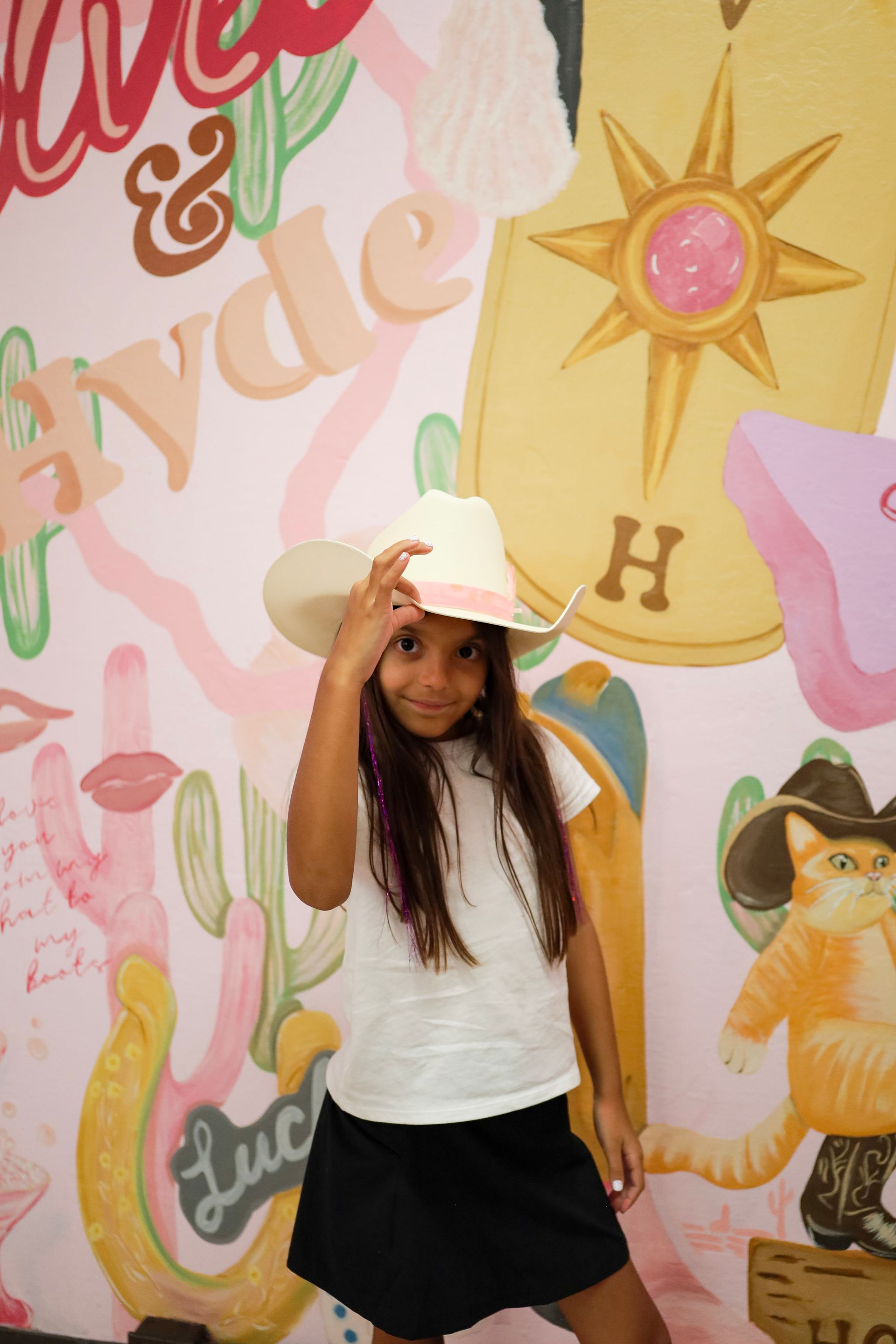 Person posing in front of a colorful mural, wearing a white shirt, black skirt, and hat.