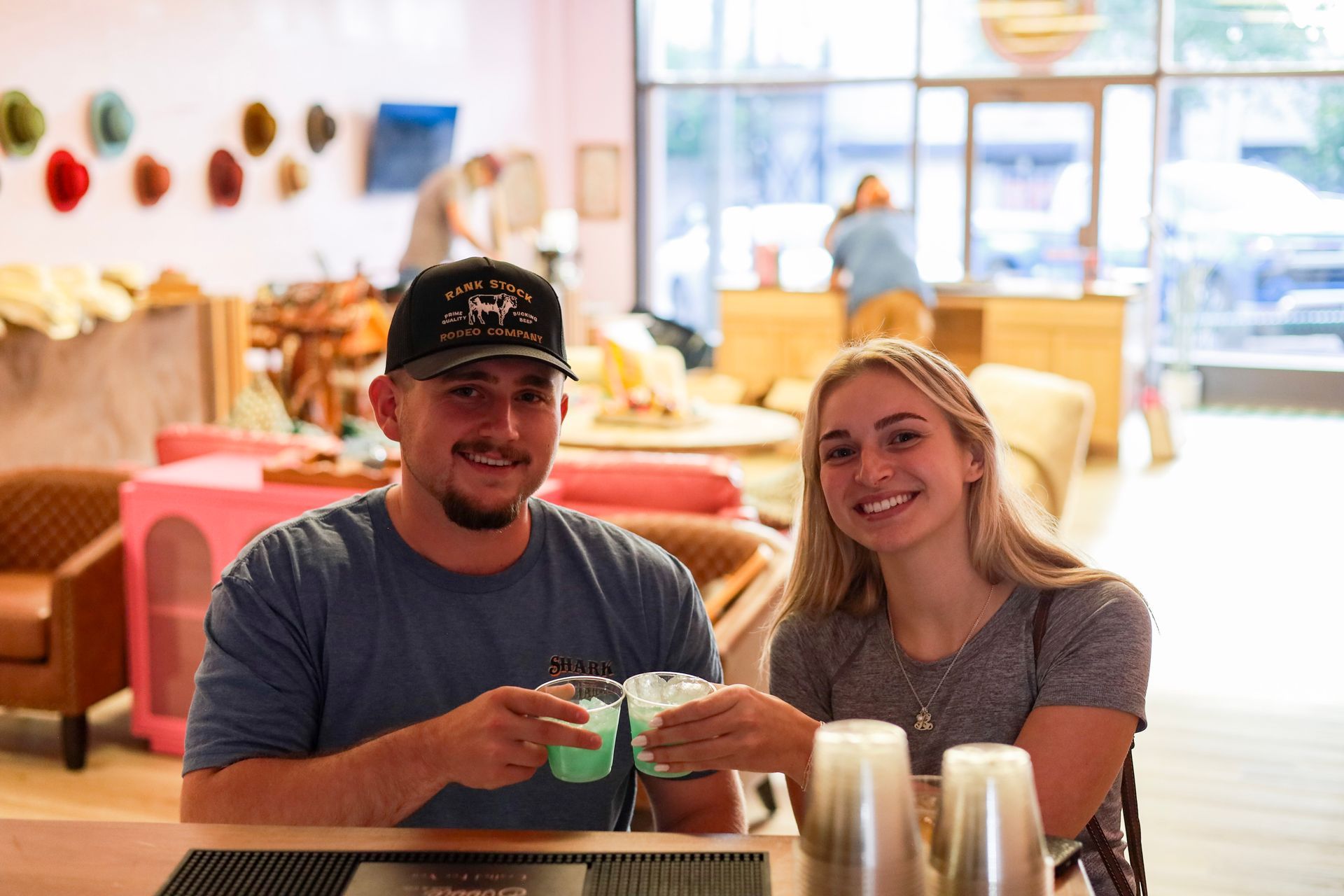 Two people smiling and clinking drinks at a cafe table, with colorful seating in the background