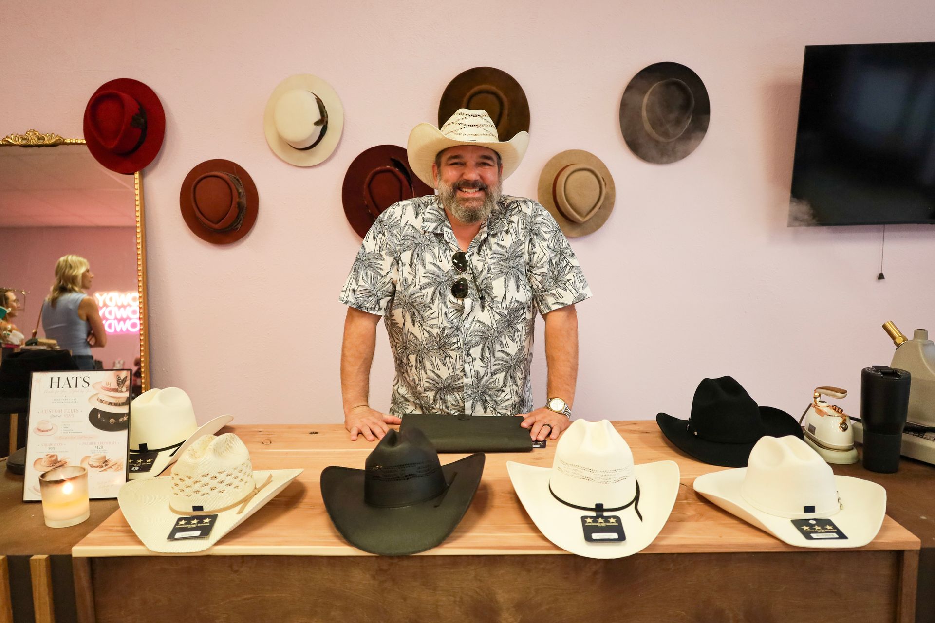 Man presenting hats on a table in a room with wall-mounted hats and a TV
