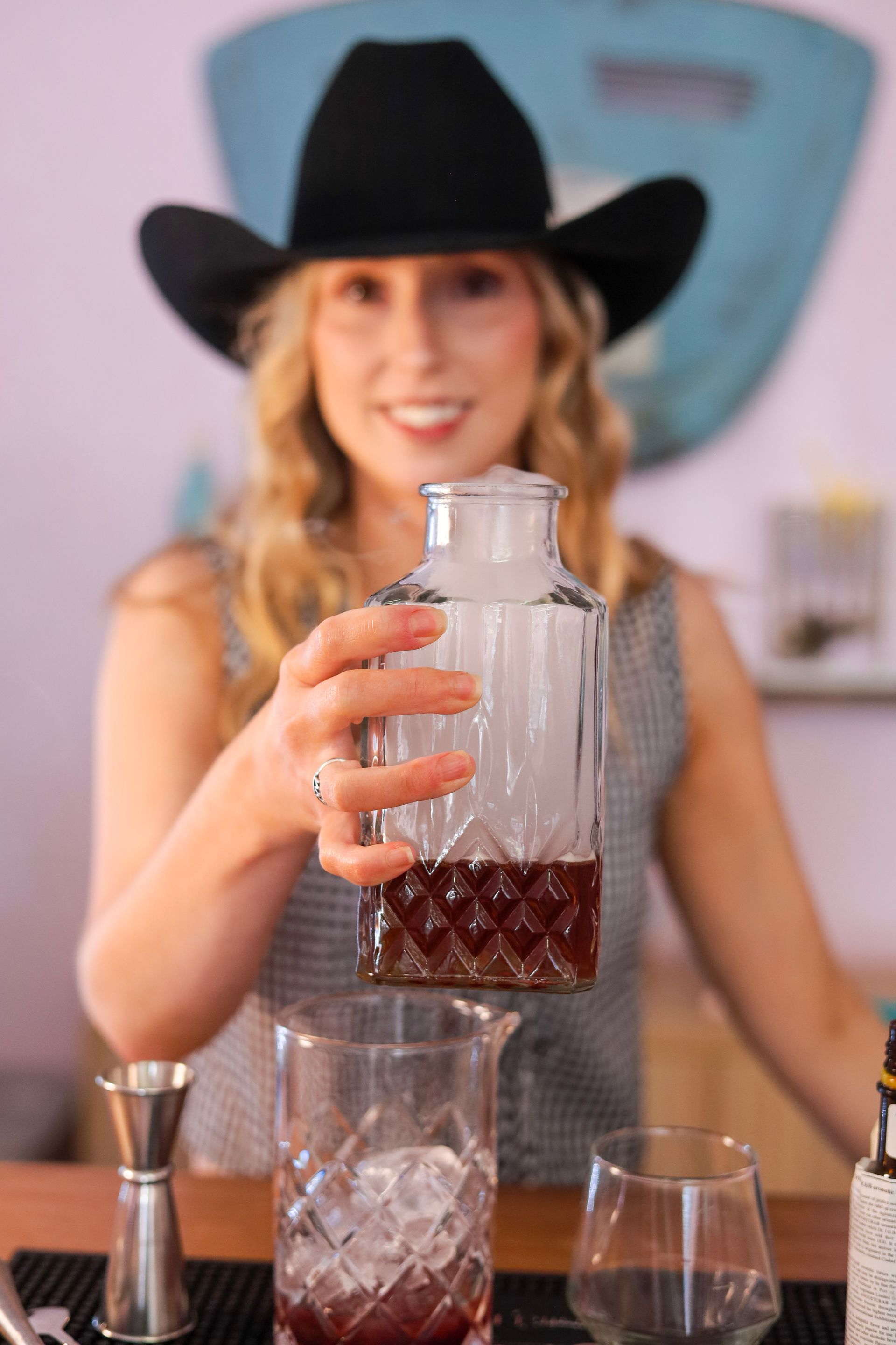 Woman in a black cowboy hat holding up a faceted glass cocktail shaker behind a bar.