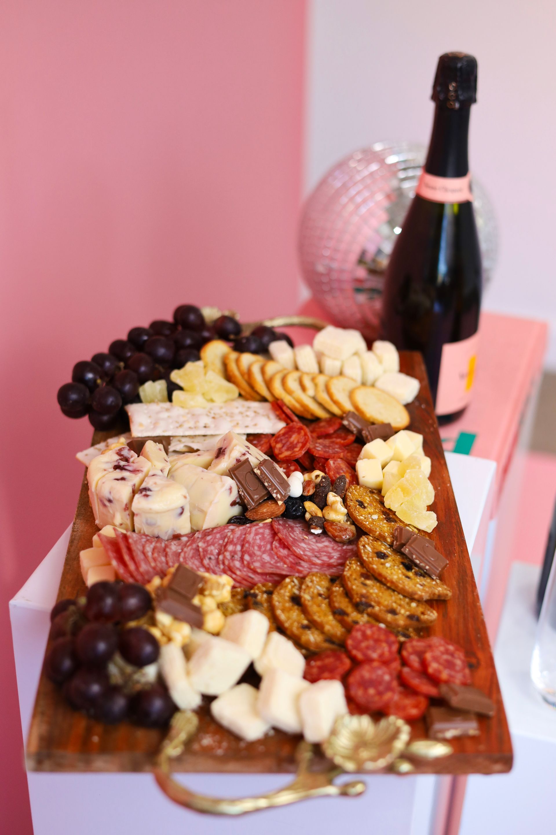 Charcuterie board with cheeses, grapes, meats, crackers, and a bottle of champagne on a pink table