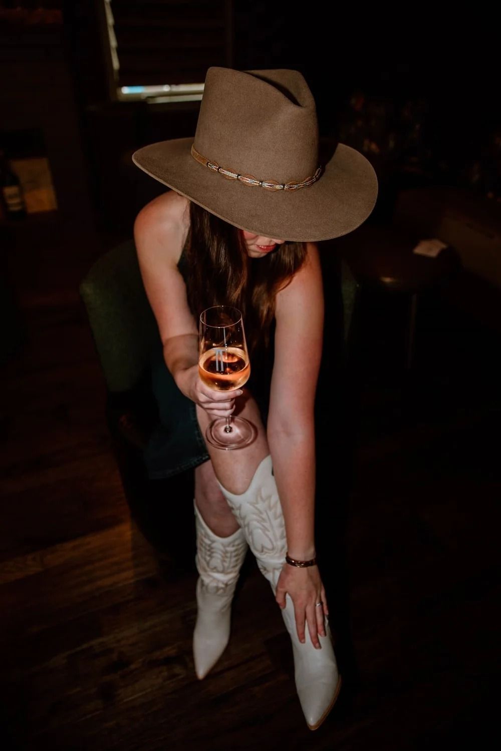 Person in tan hat and white boots crouches indoors, holding a drink with one leg crossed over the other.