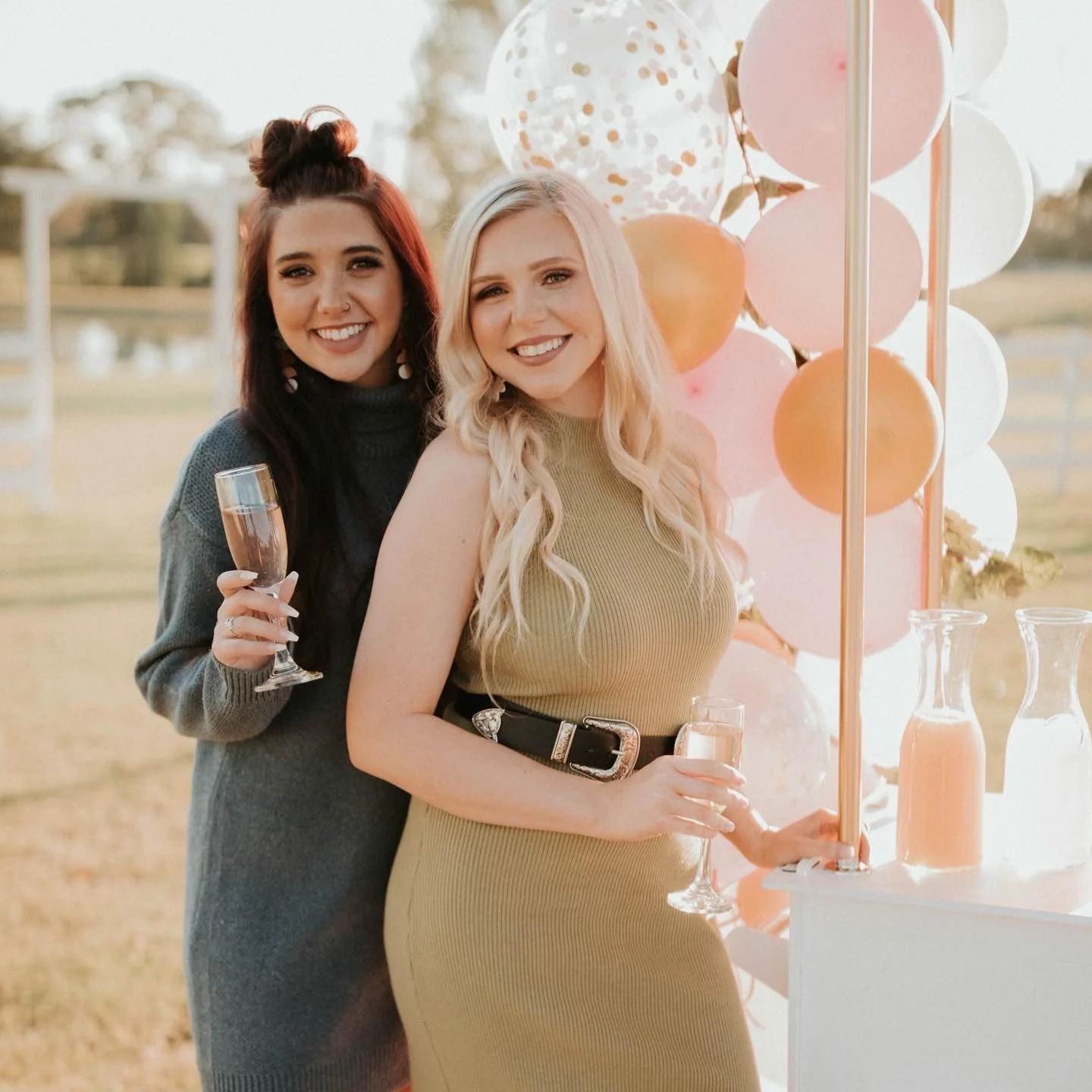 Two women smiling beside a pastel balloon display at an outdoor party, one holding a drink