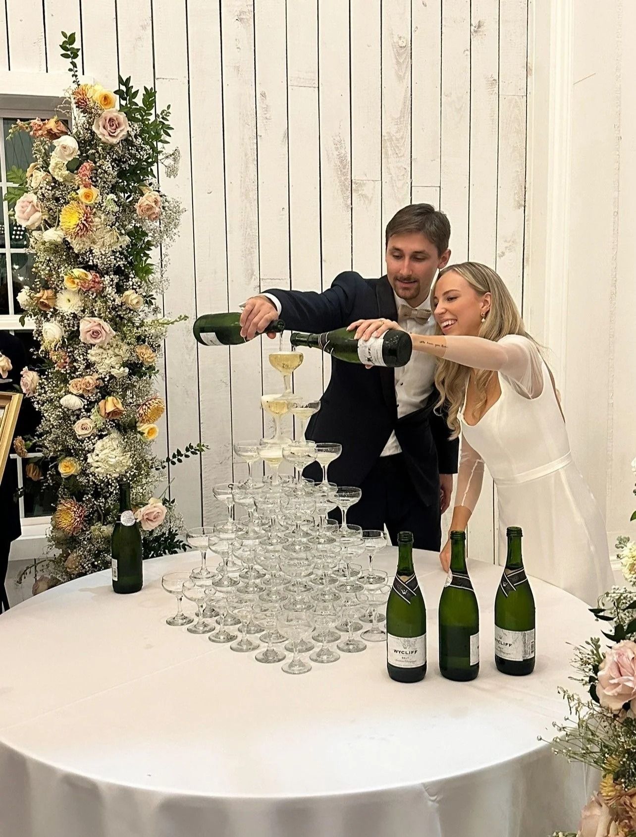 Bride and groom pour champagne into a tower of glasses at a floral wedding reception table