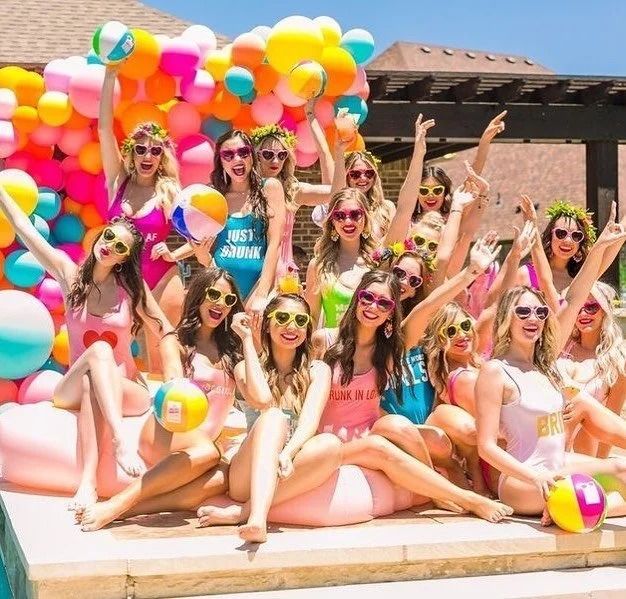 Group posing with colorful balloons in a sunny outdoor poolside setting, smiling and wearing sunglasses.