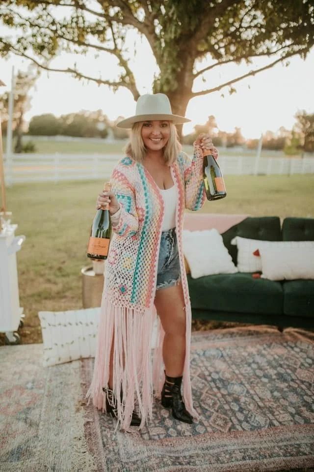 Woman in colorful fringe outfit holding a bottle outdoors beside a tree and couch