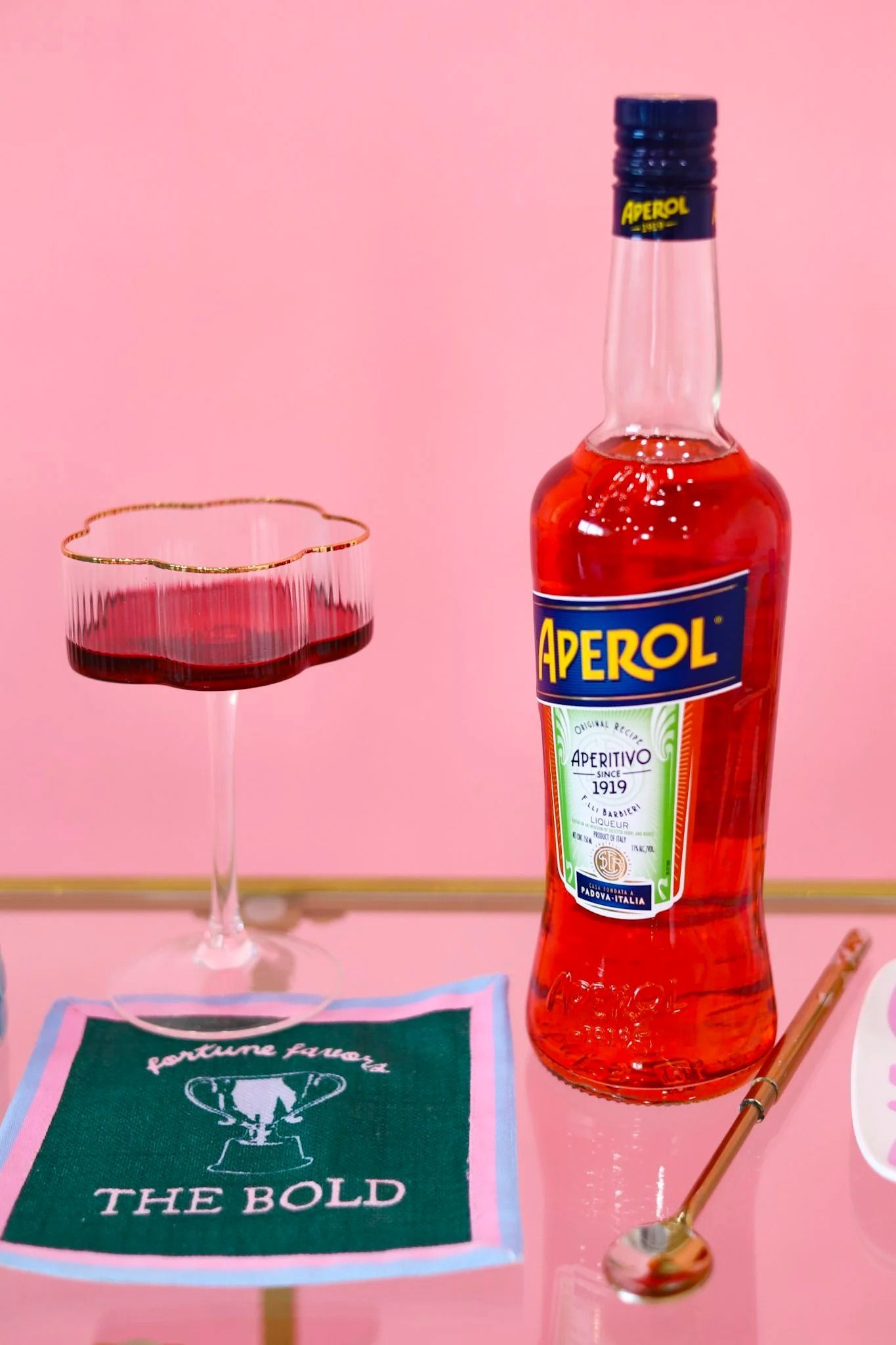 Aperol bottle and cocktail glass on a pink table with a green napkin and gold spoon.