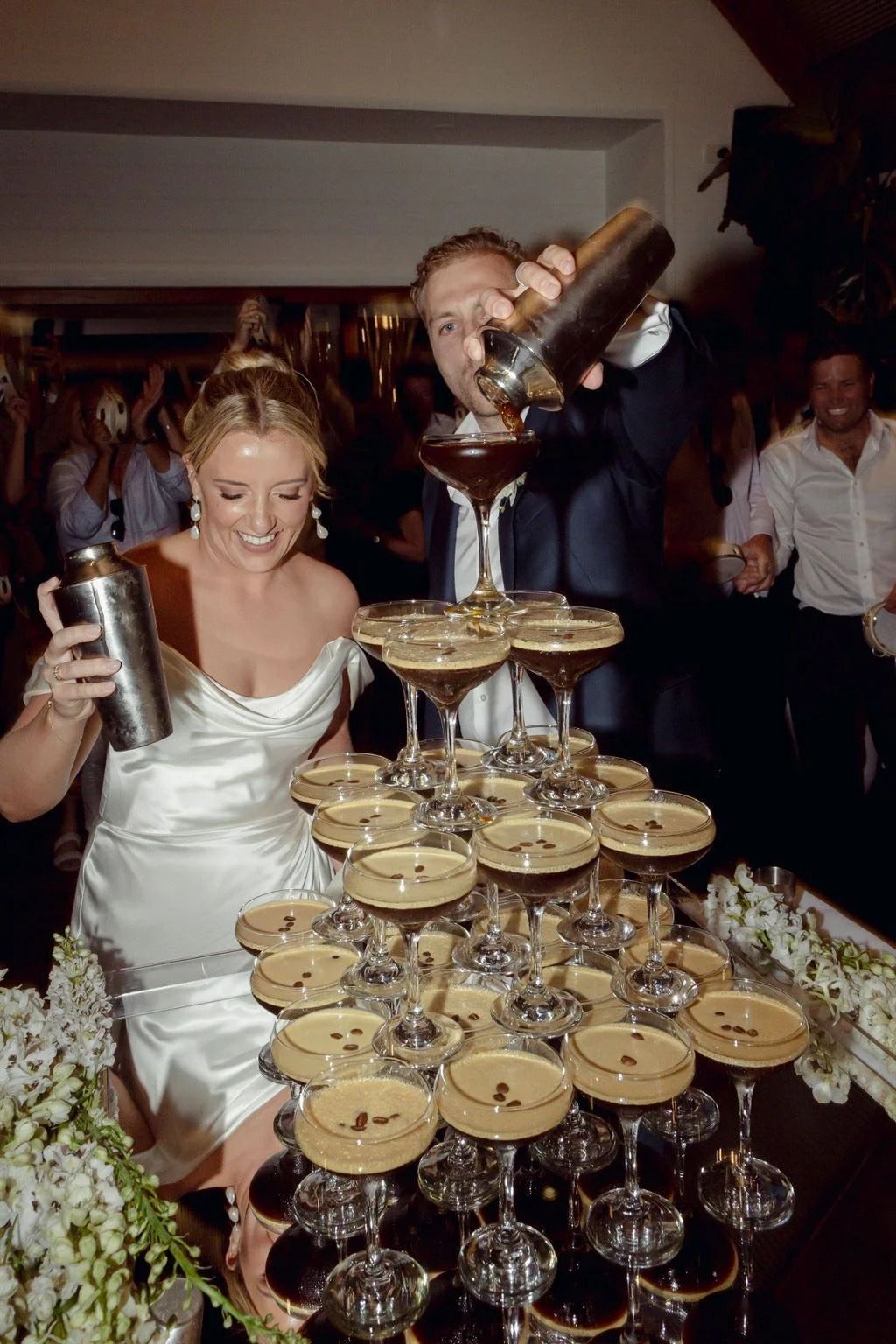 Bartender stacking cocktails into a champagne tower while a woman in a white dress smiles nearby